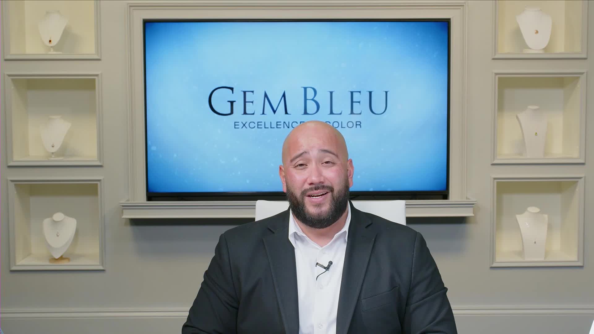 A man in a suit, speaking animatedly, sits in front of a screen with the "Gem Bleu" logo.  Jewelry displays are arranged on the wall behind him, likely part of a live broadcast from the Gem Shopping Network.
