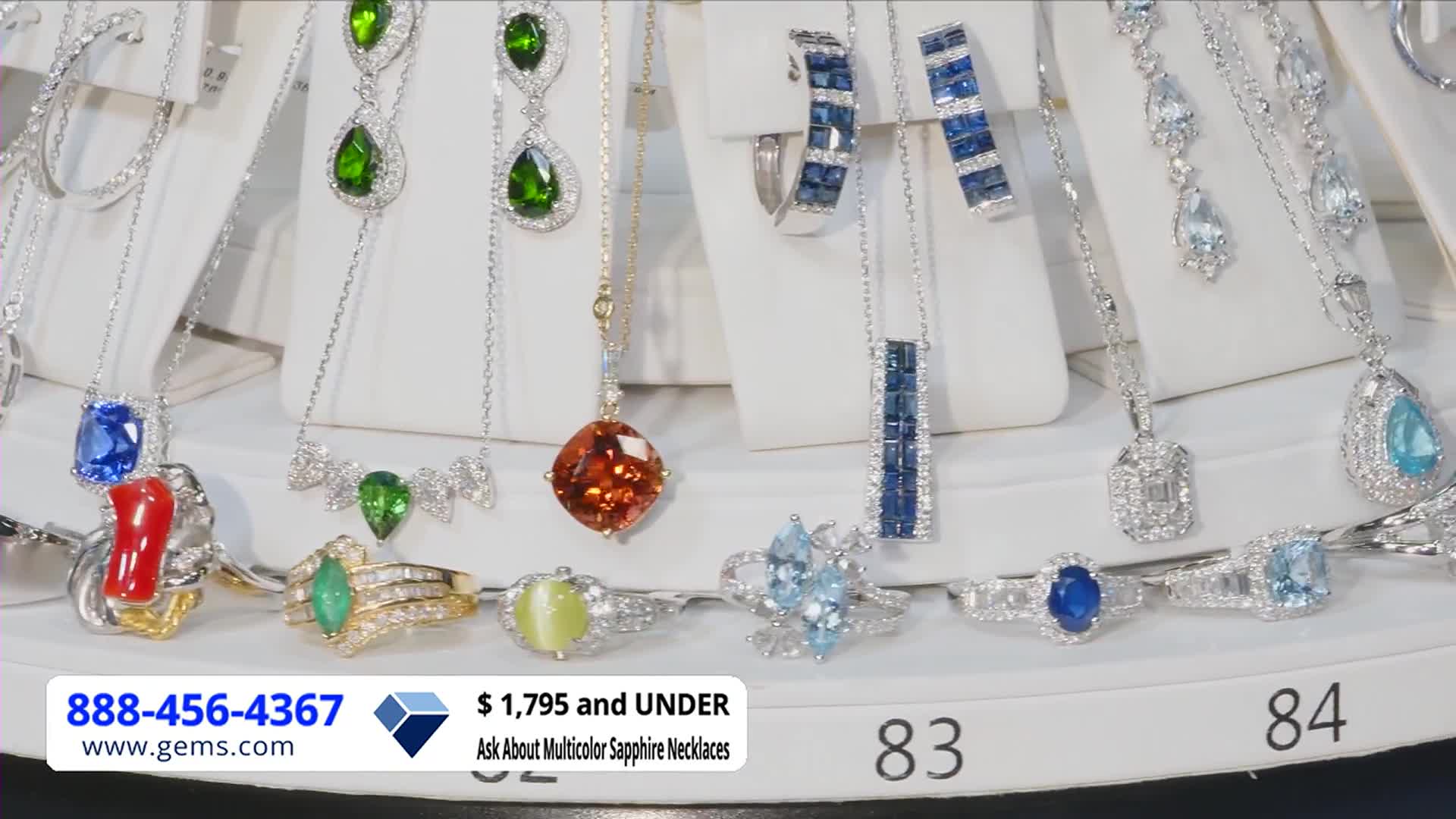 A dazzling array of necklaces and rings sparkle under bright lights, all displayed on white stands. The Gem Shopping Network is advertising pieces for $1,795 and under, with a phone number and website displayed.
