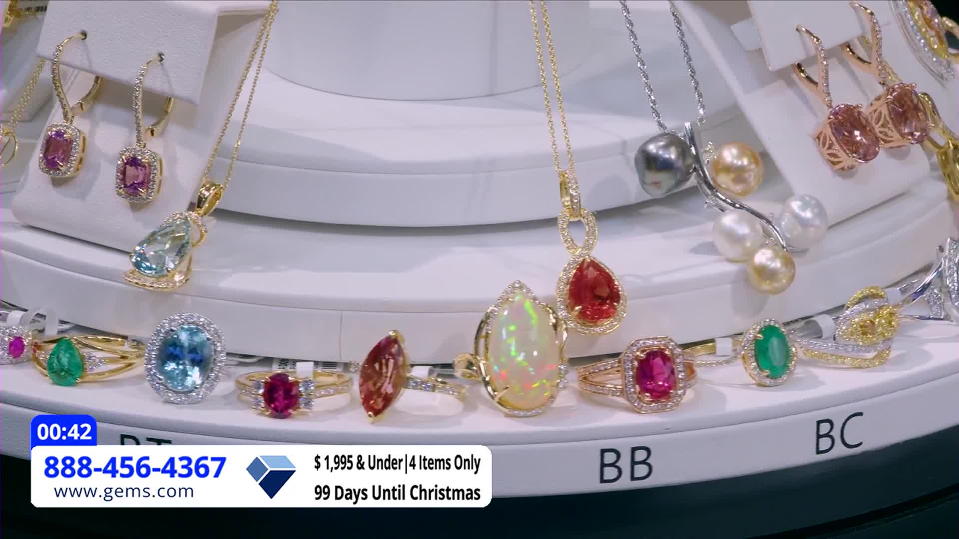 A rotating display showcases rings, necklaces, and earrings from the Gem Shopping Network, all sparkling under bright lights. The screen displays a phone number and website, along with a countdown to Christmas.
