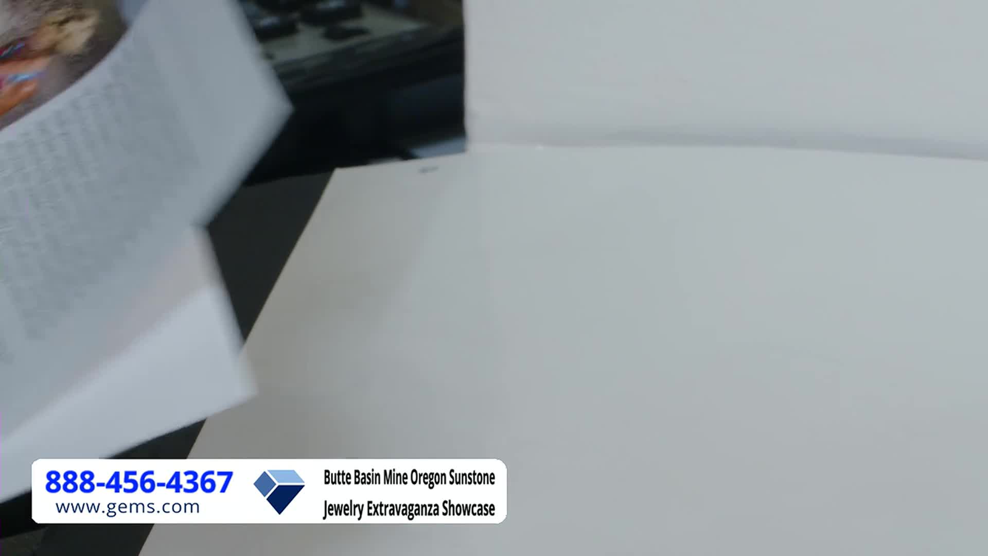 A hand flips through pages of a document, likely a script for the Gem Shopping Network. The camera angle focuses on the white paper, with the Butte Basin Mine Oregon Sunstone Jewelry Extravaganza Showcase banner visible in the corner.
