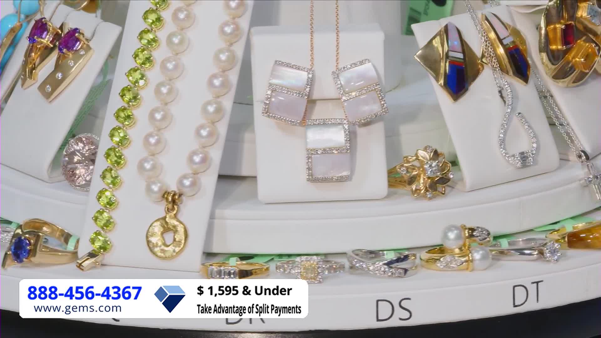 A display of jewelry sparkles under bright lights, with necklaces, rings, and earrings arranged on white stands. The Gem Shopping Network logo and a phone number are visible at the bottom of the screen.
