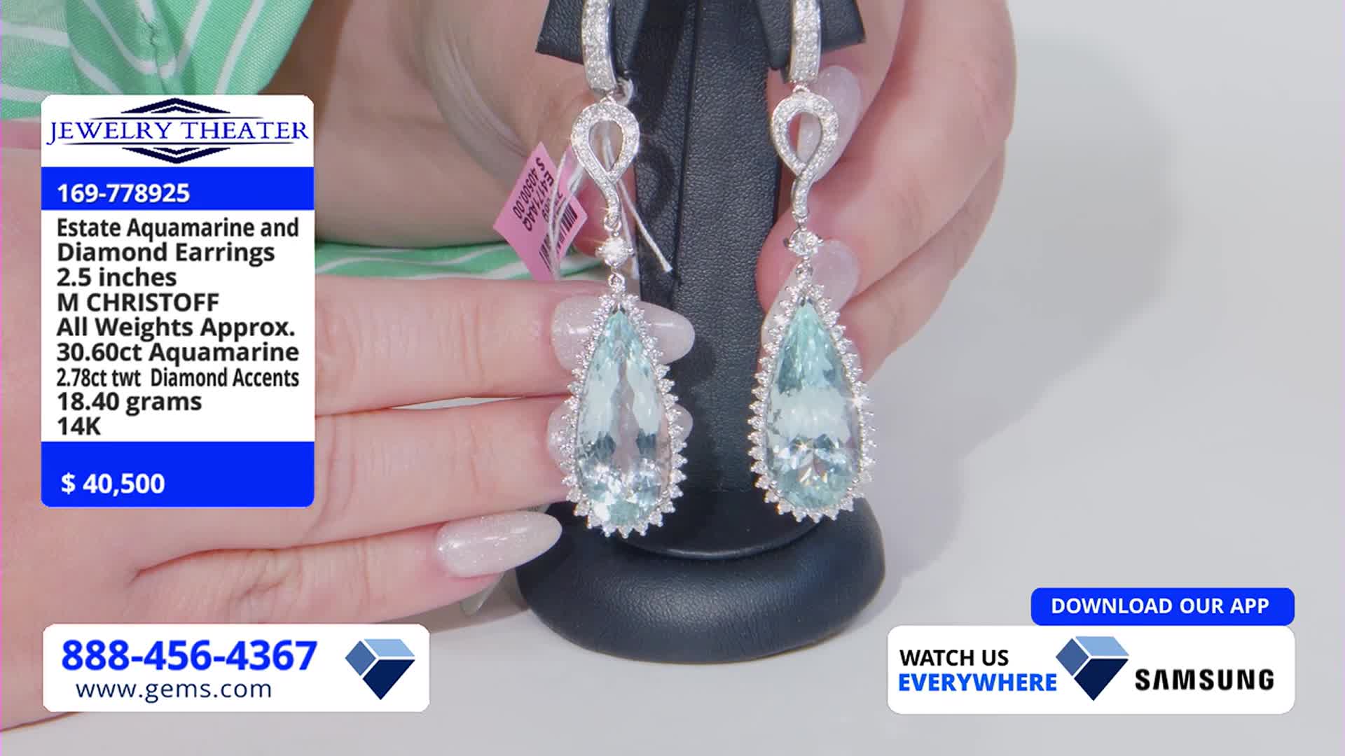 A pair of aquamarine and diamond earrings hangs from a small stand, held up by manicured hands. The Gem Shopping Network's price tag dangles near the top of one earring.
