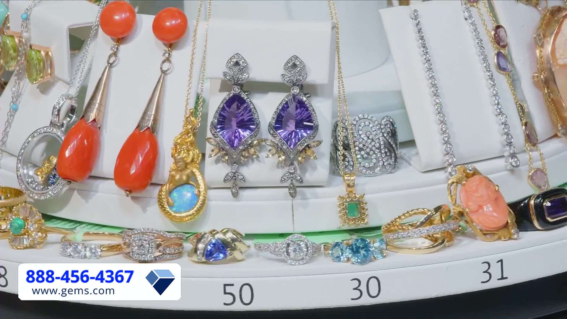 A dazzling array of jewelry, from earrings with large purple stones to coral pendants, is displayed on a revolving platform. The Gem Shopping Network's phone number and website are prominently visible in the lower left corner.
