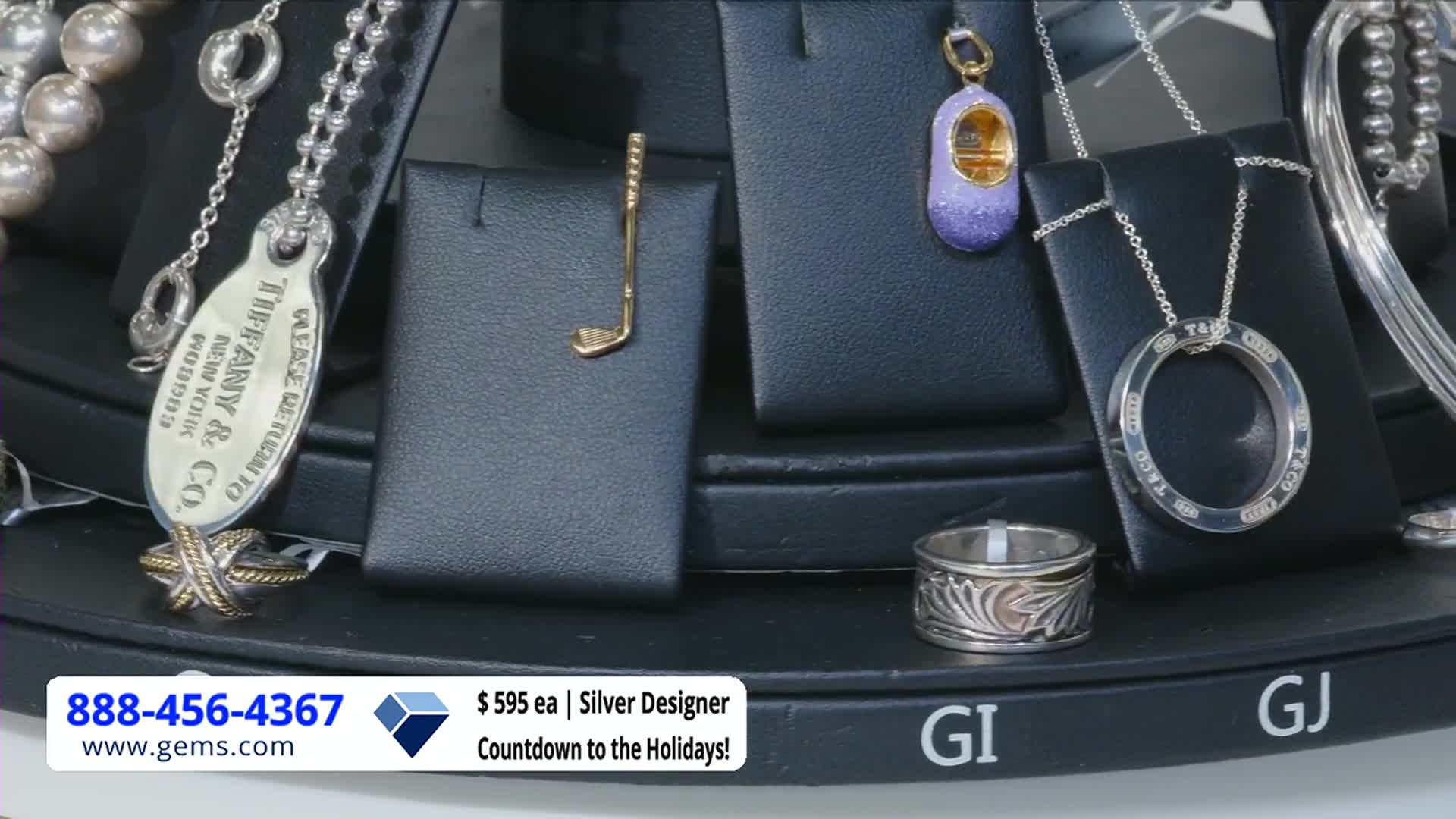 A rotating display of jewelry is set up, including a golf club pendant and a ring with an engraved pattern. The Gem Shopping Network logo and a phone number are visible at the bottom.

