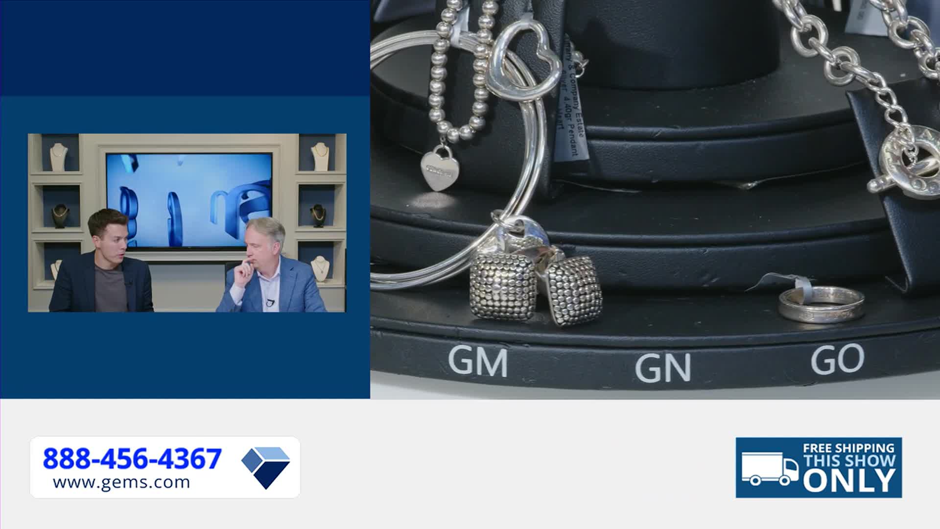 Two men on a Gem Shopping Network set discuss jewelry, while a rotating display shows off the pieces. The display features silver necklaces, earrings, and a ring.
