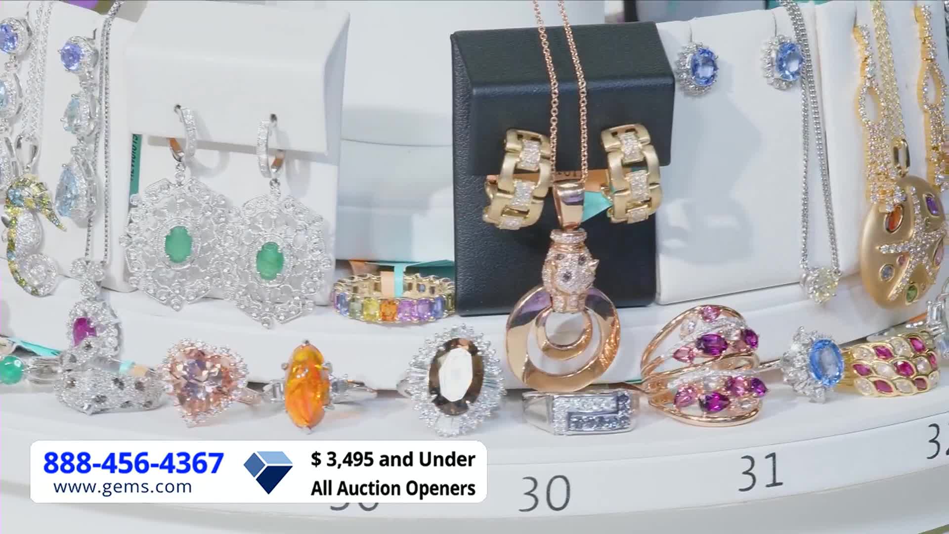 On a Gem Shopping Network set, jewelry is displayed on tiered platforms.  Rings, necklaces, and earrings sparkle under bright lights, each piece ready to be presented.
