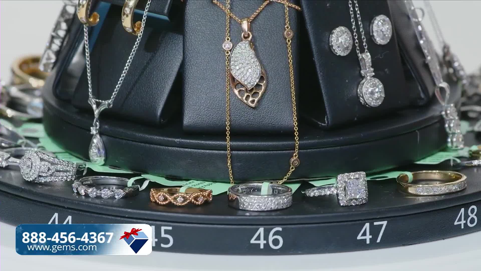 Jewelry, rings and necklaces, are displayed on a rotating platform, with prices and item numbers visible. A Gem Shopping Network logo and phone number are in the lower left corner.
