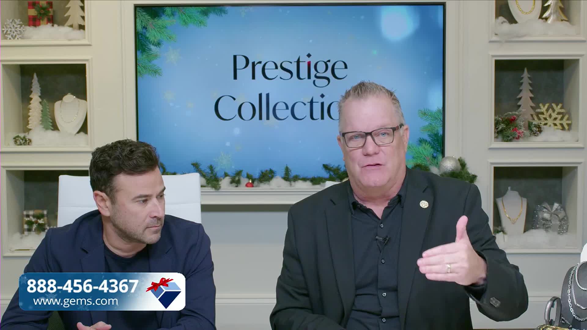 A man in a black suit gestures with his right hand, speaking to the camera. His colleague sits beside him, listening intently.
