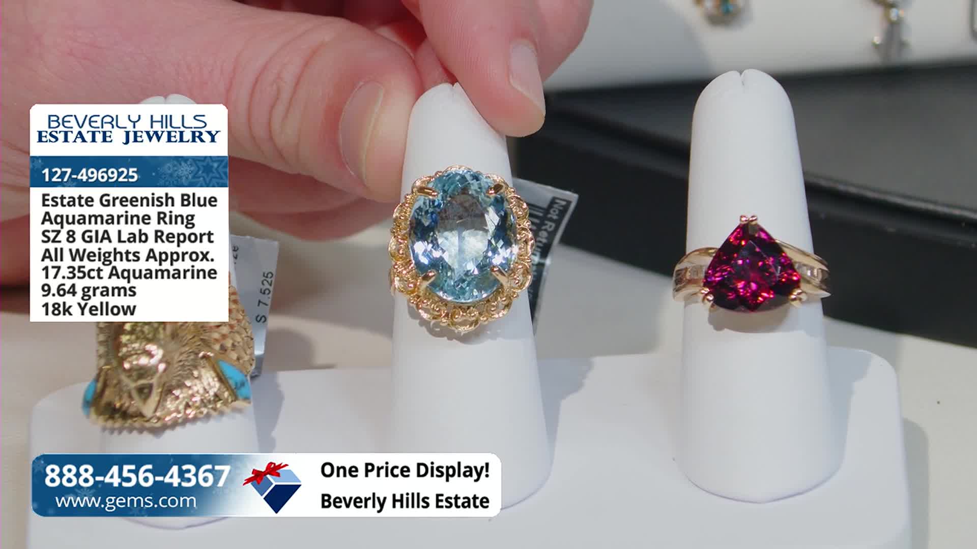 A hand presents a large, oval aquamarine ring, its light blue stone catching the light. Beside it, another ring with a triangular, deep red stone sits on a white display.
