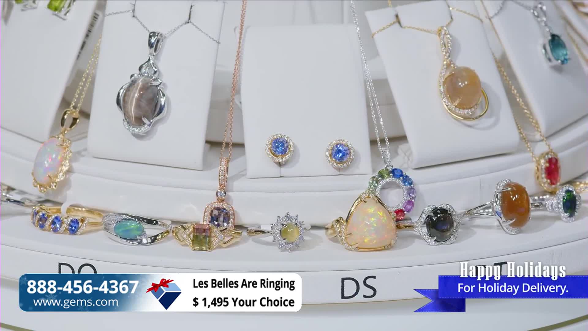 On a white display, necklaces and rings with colorful gemstones are arranged on a Gem Shopping Network set. The camera focuses on the jewelry, with a phone number and website address visible at the bottom of the screen.
