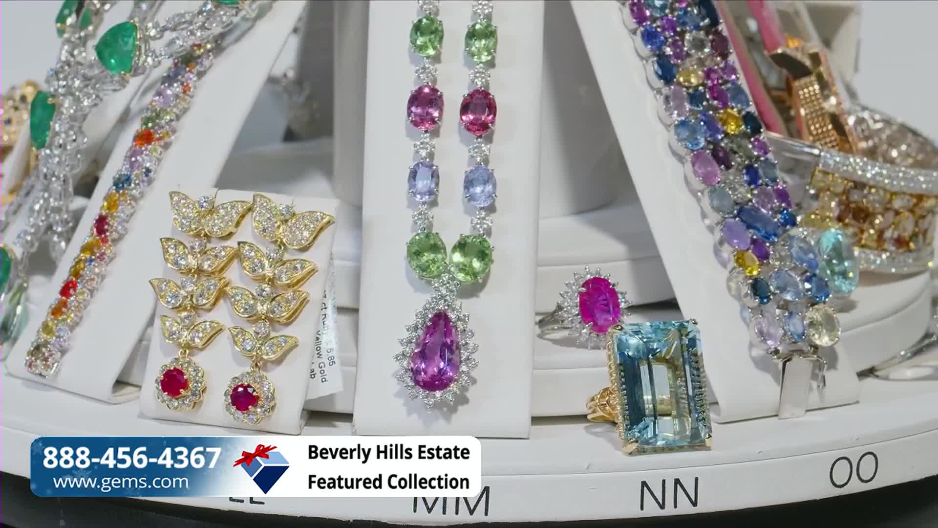 Jewelry, including necklaces and rings, is displayed on white pedestals, each piece catching the light. The Gem Shopping Network logo and a phone number are visible in the lower left corner.
