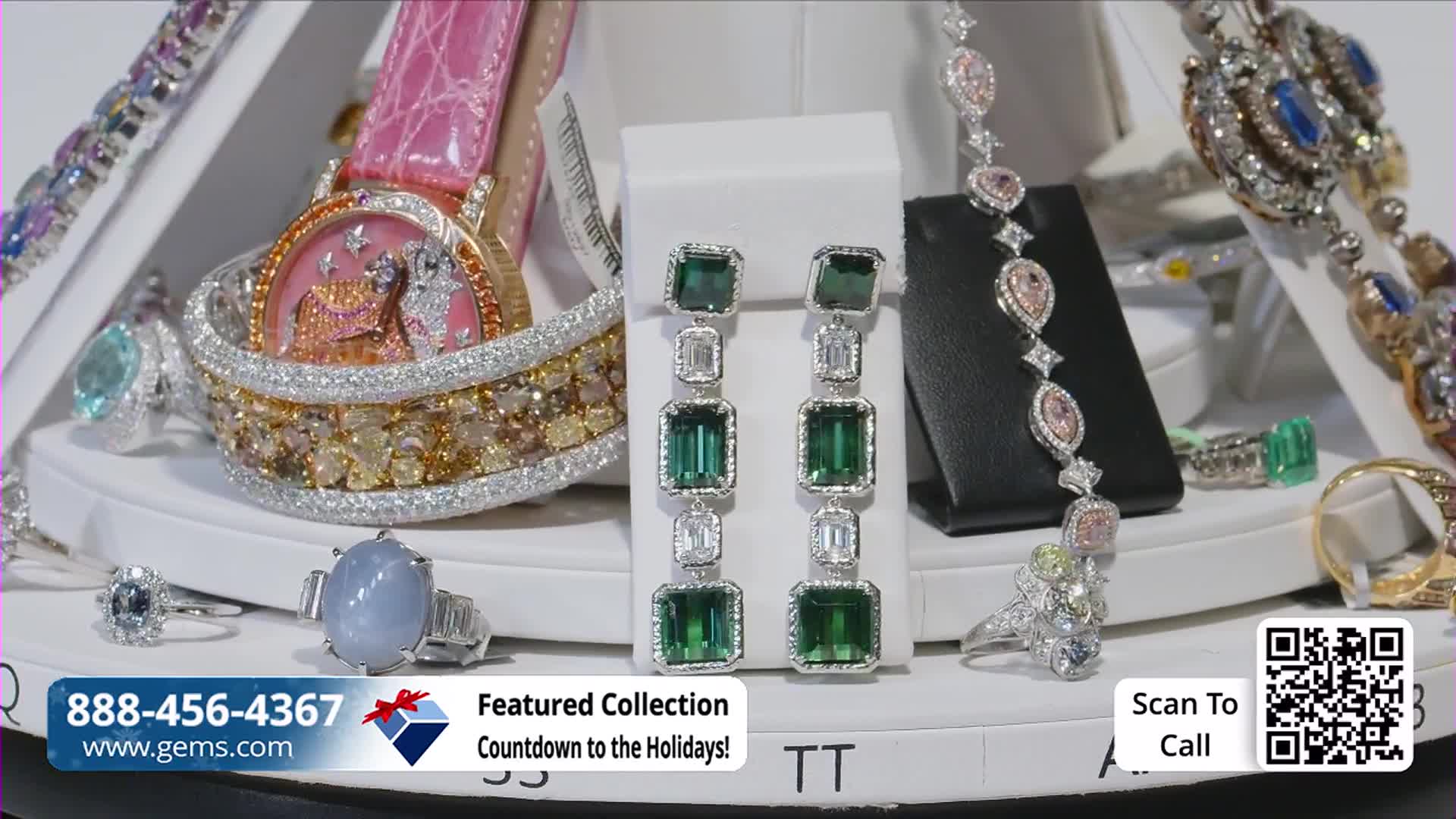 A display of jewelry sparkles under bright lights, with long emerald earrings front and center. A pink-strapped watch and various rings and bracelets are arranged around the earrings, all ready to be featured on Gem Shopping Network.
