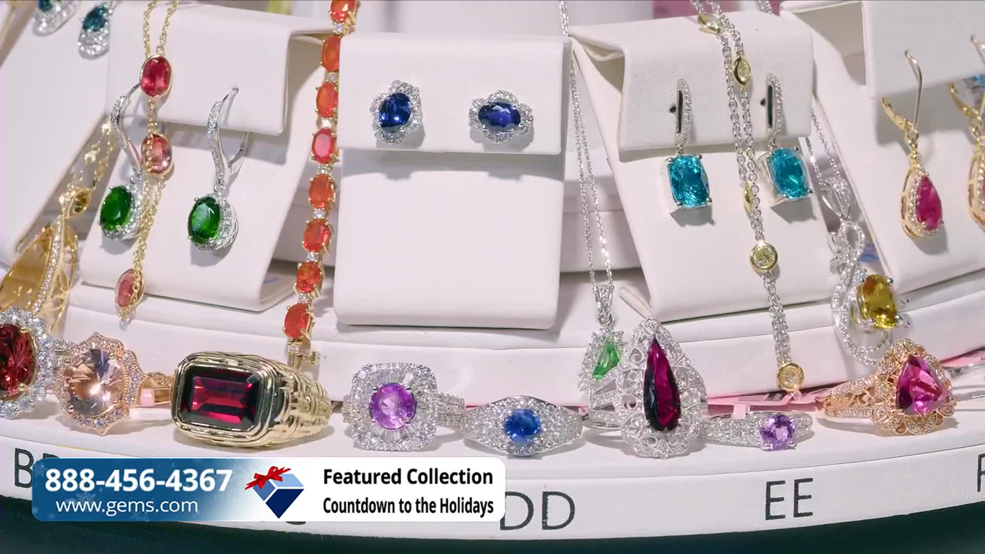 Jewelry, displayed on white pedestals, sparkles under bright lights. The Gem Shopping Network logo and a phone number are visible at the bottom of the screen.

