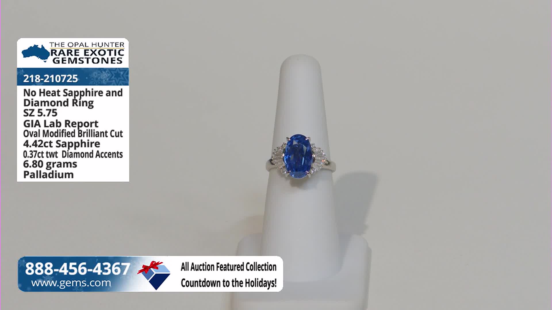 A sapphire ring, set on a white display, is centered against a plain background. The ring's large, oval blue stone is flanked by smaller, clear accent stones.
