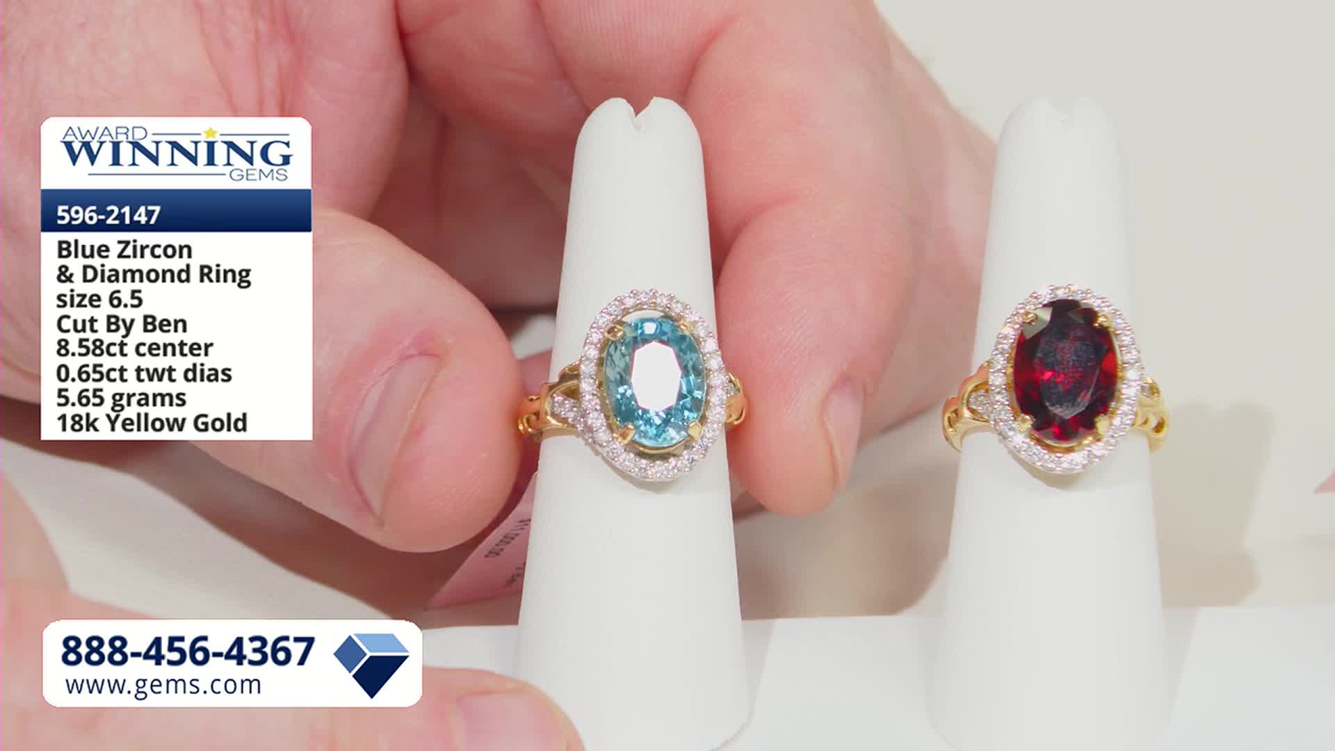 A hand turns a ring with a striking blue zircon, surrounded by small diamonds, on a white display. Another ring, featuring a deep red stone and similar diamond halo, sits beside it.