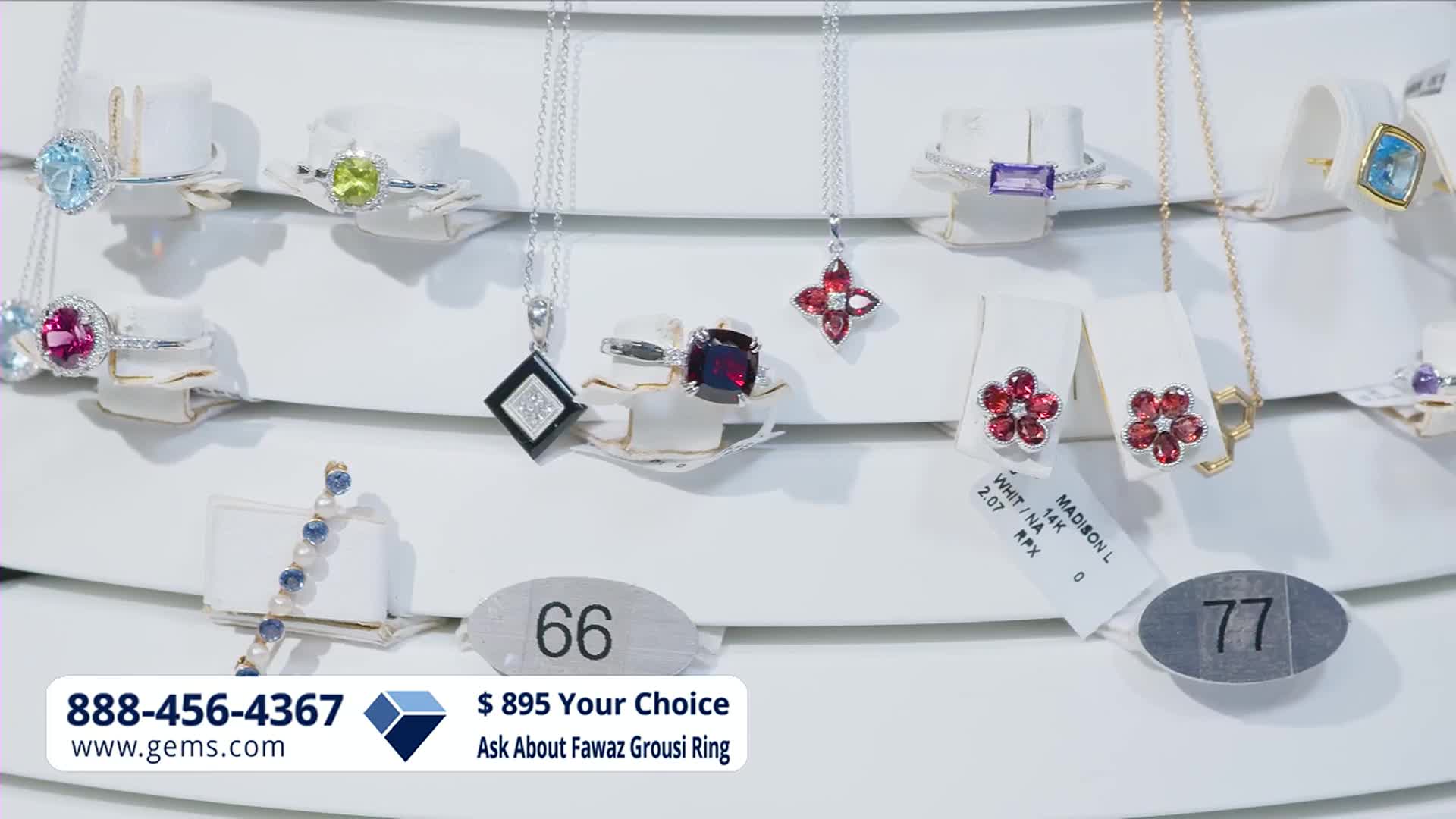 A large, square black pendant with a diamond inlay hangs from a delicate silver chain. To its right, a ring with a deep red gemstone sits on a white display.