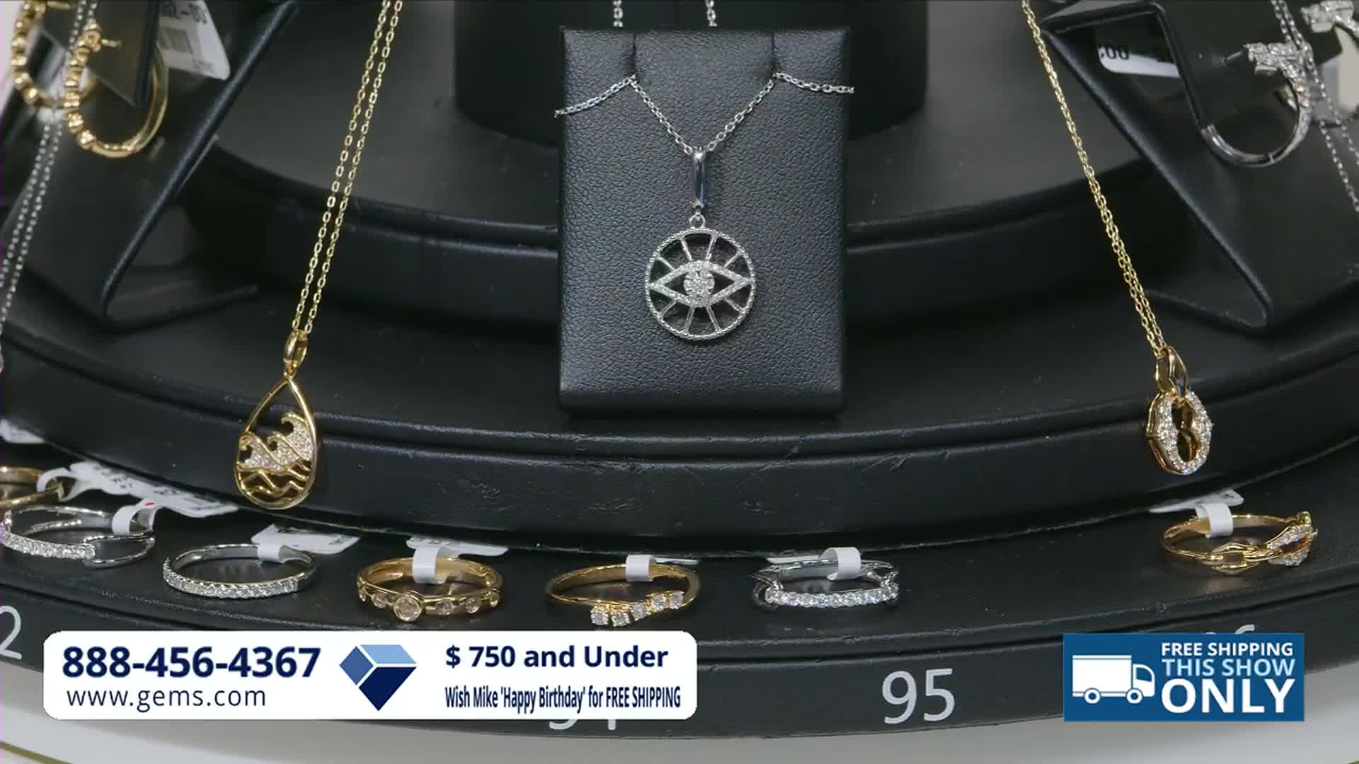 A gold pendant with a wave design hangs from a delicate chain. Below, several diamond-studded rings are displayed on a black tiered stand.