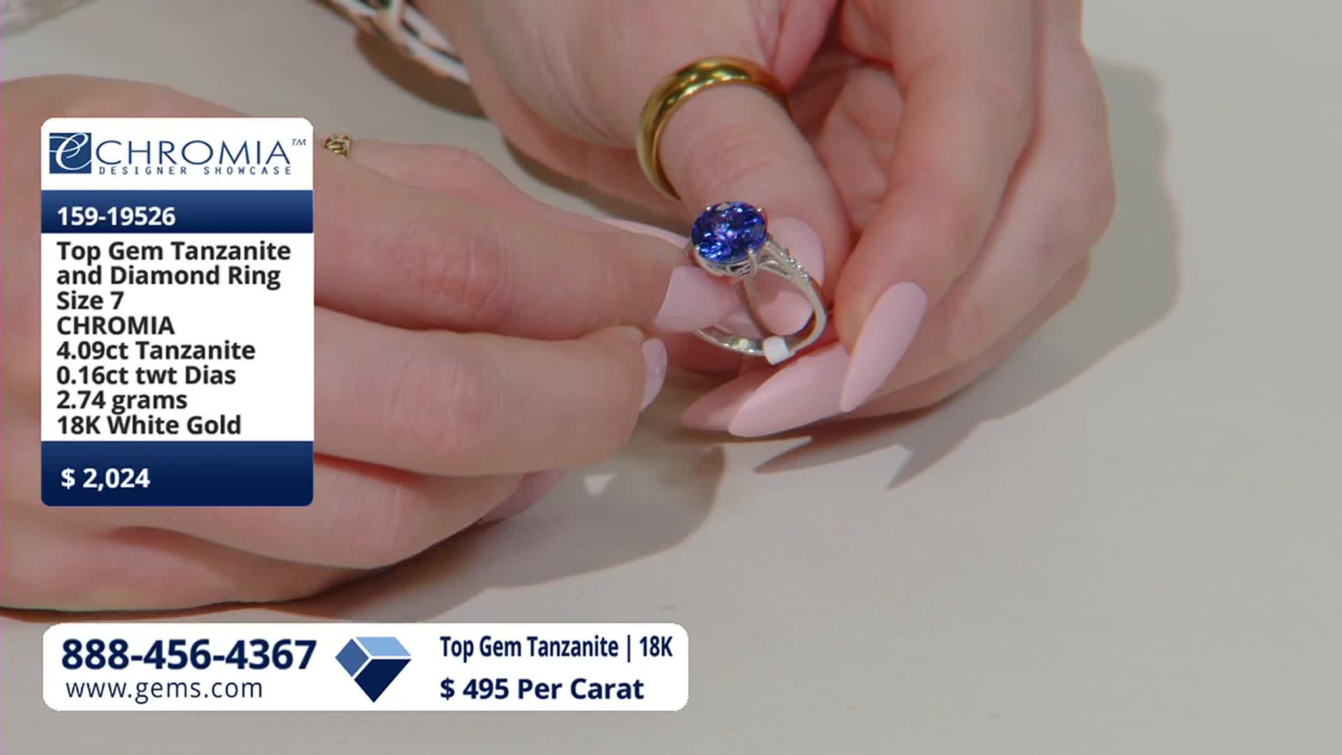 A hand with pale pink nails turns a silver ring, showcasing a large, deep blue tanzanite stone. The ring is being presented on the Gem Shopping Network.