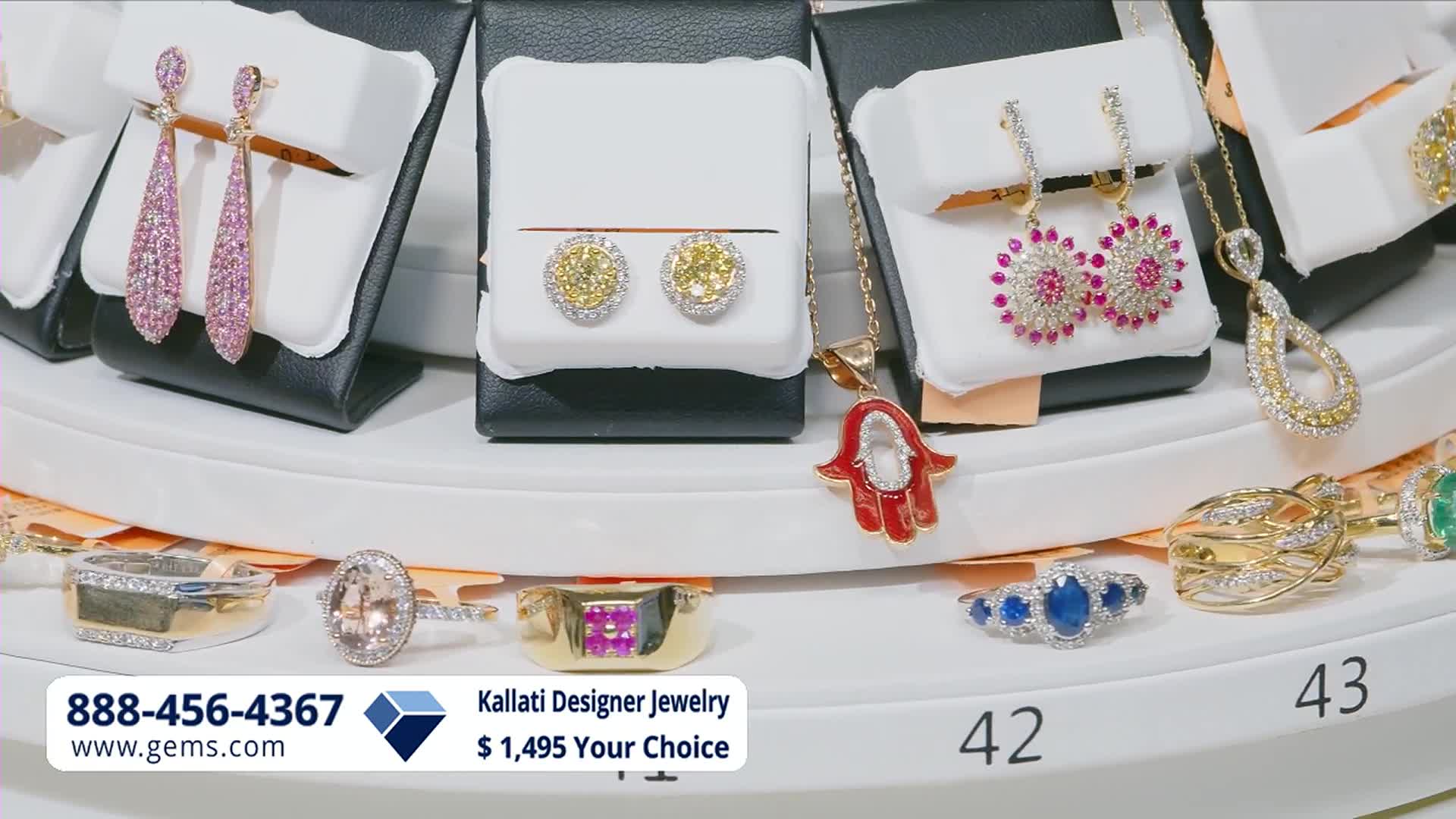 Sparkling jewelry, including dangling pink sapphire earrings and yellow diamond studs, are displayed on white stands. Rings and pendants, featuring colorful gemstones and intricate gold settings, rest on a tiered display.