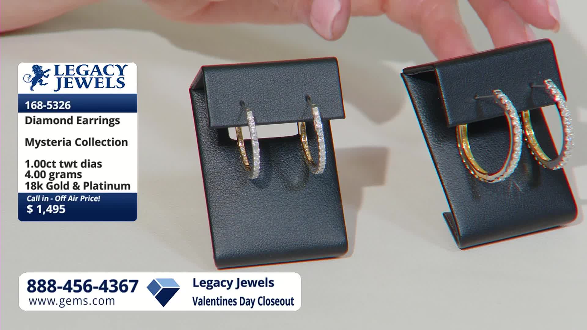 A hand reaches out to adjust a pair of diamond hoop earrings on display. Another set of similar earrings sits nearby, also on a black stand.