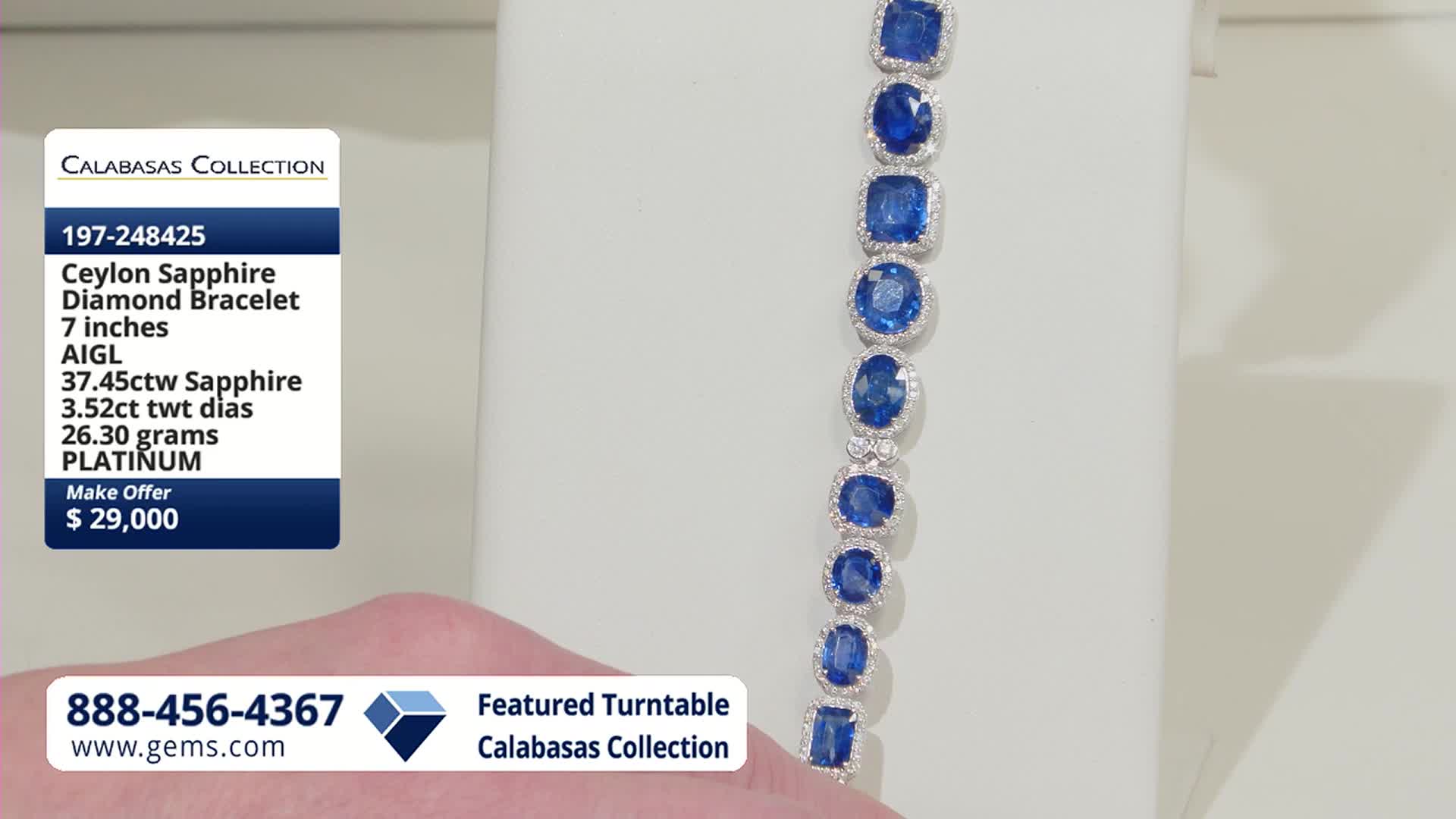 A platinum bracelet with large, deep blue sapphires, each surrounded by tiny diamonds, rests on a white stand. The Gem Shopping Network's phone number and website are displayed at the bottom.