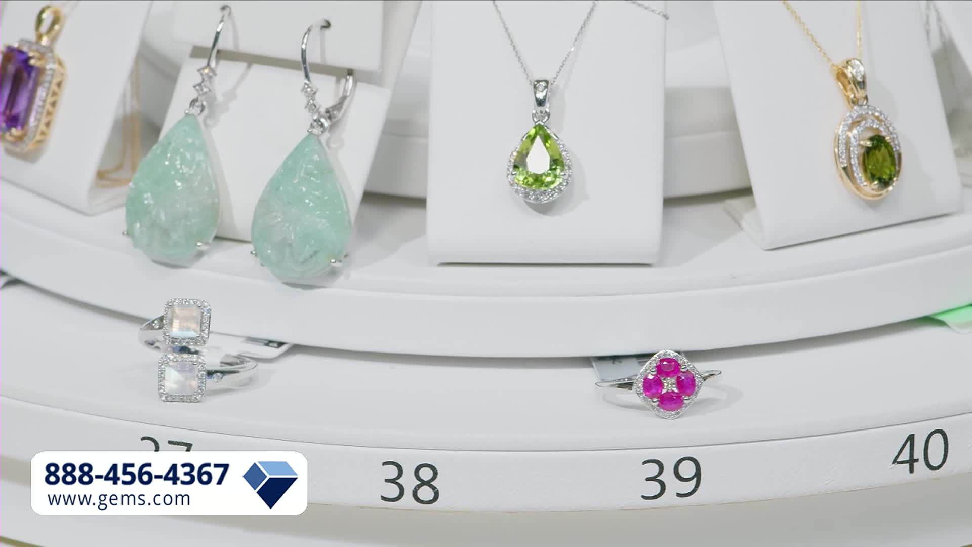 A display of jewelry rotates, showcasing pendants and earrings with colorful gemstones. Two rings, one with stacked square stones and another with a floral ruby design, sit on the lower tier.
