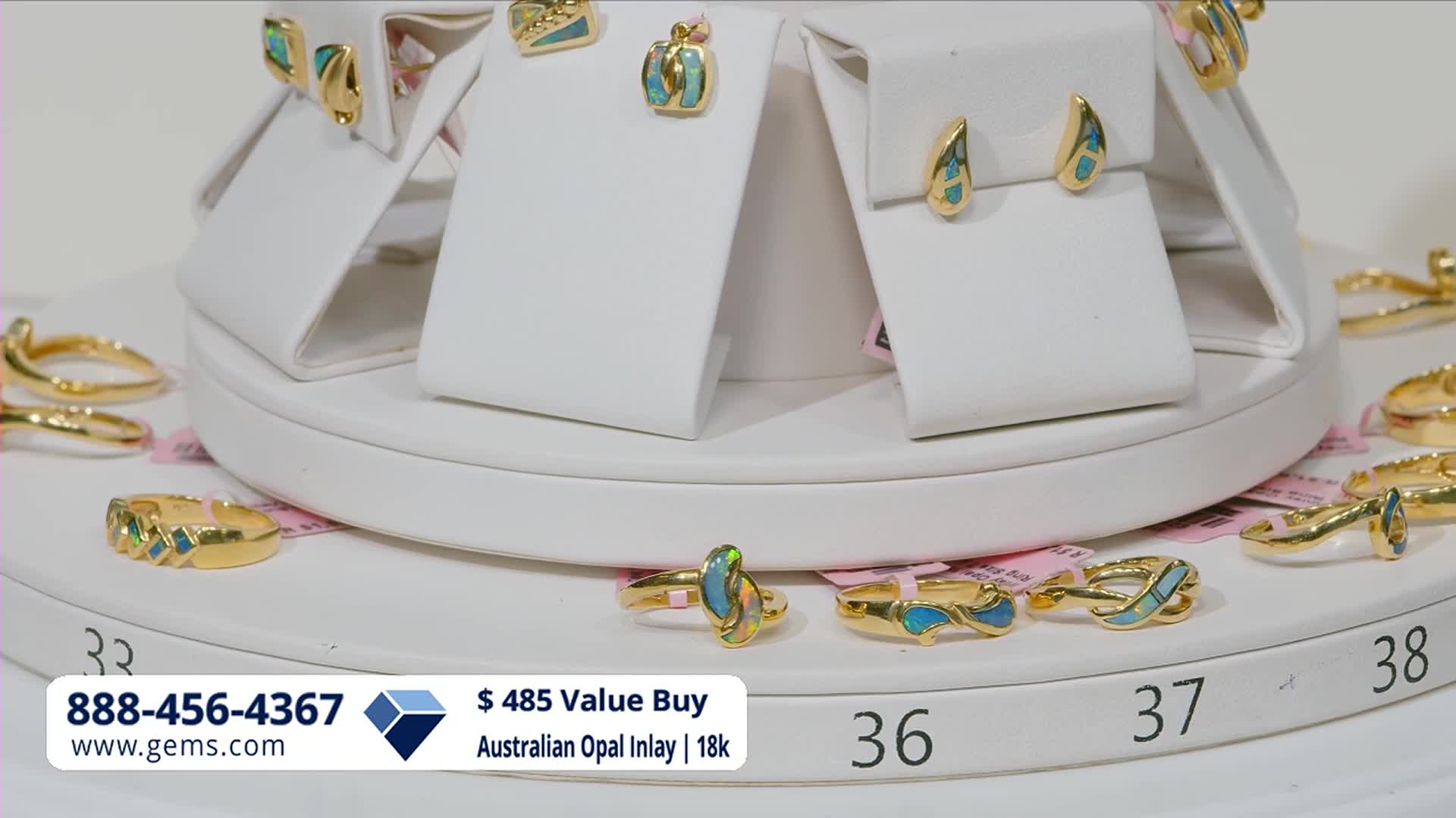 A display of gold jewelry with opal inlay sits on a white tiered stand. Rings and earrings gleam under the lights, some featuring vibrant blue and green stones.