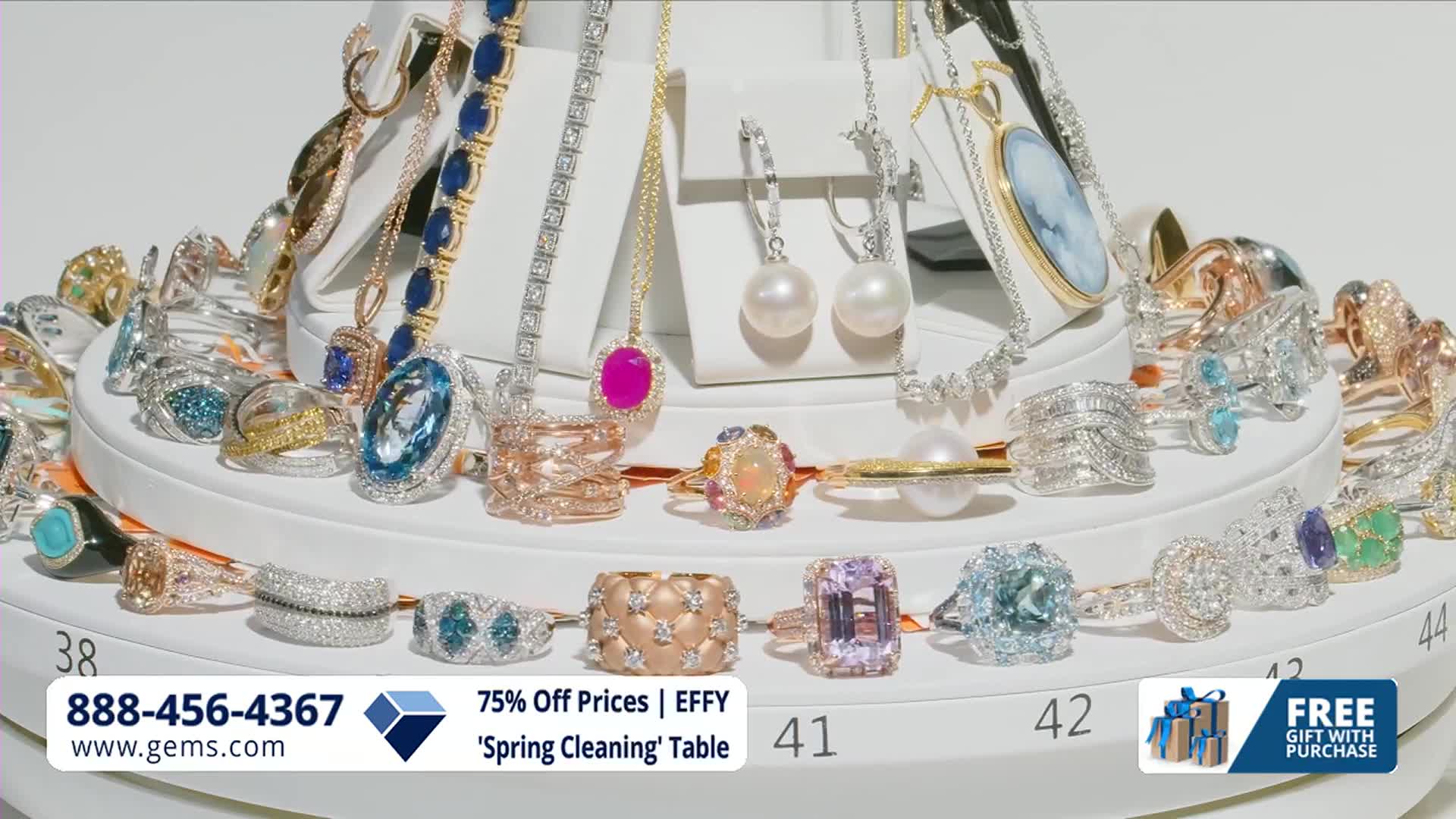 A dazzling array of rings, necklaces, and earrings are displayed on tiered stands. The Gem Shopping Network is promoting a "Spring Cleaning" sale with special prices.
