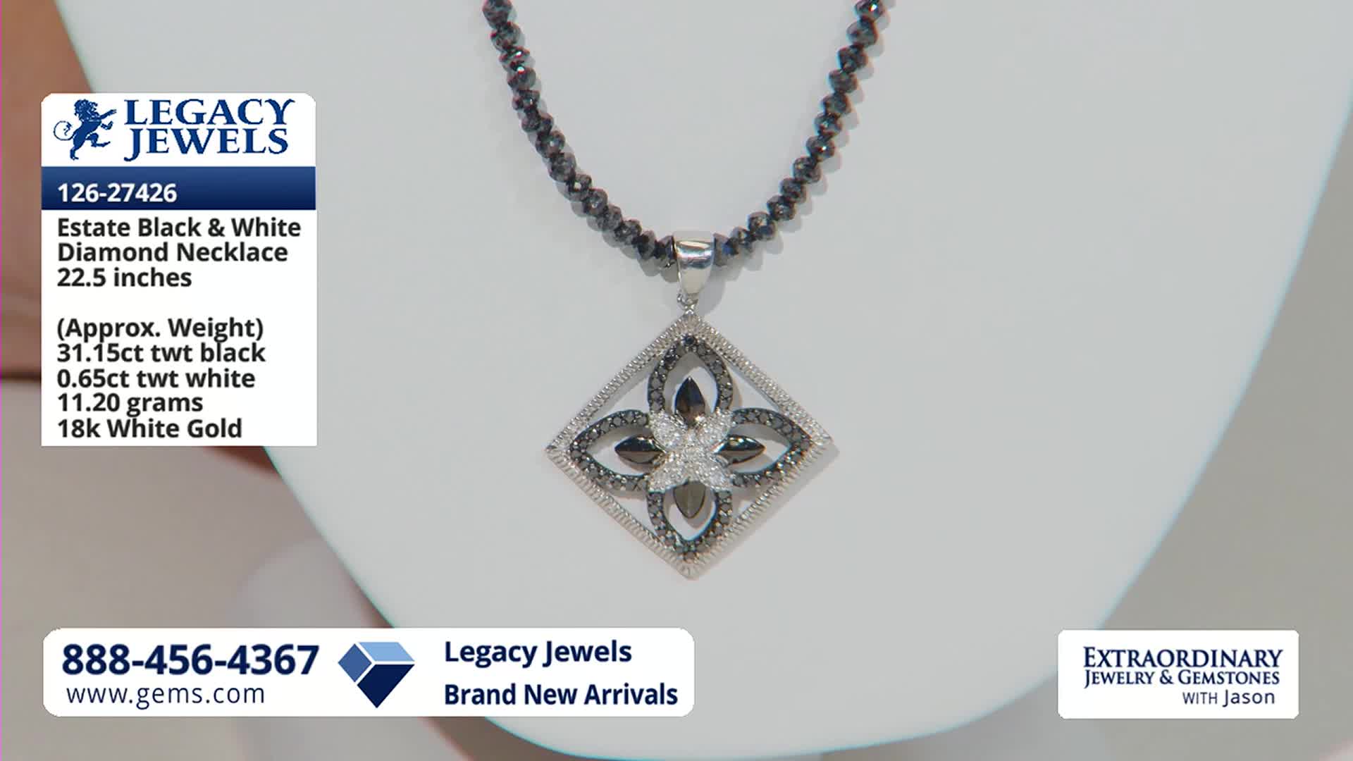 A necklace with a diamond pendant hangs on a white display. The pendant features a floral design with black and white diamonds set in 18k white gold.