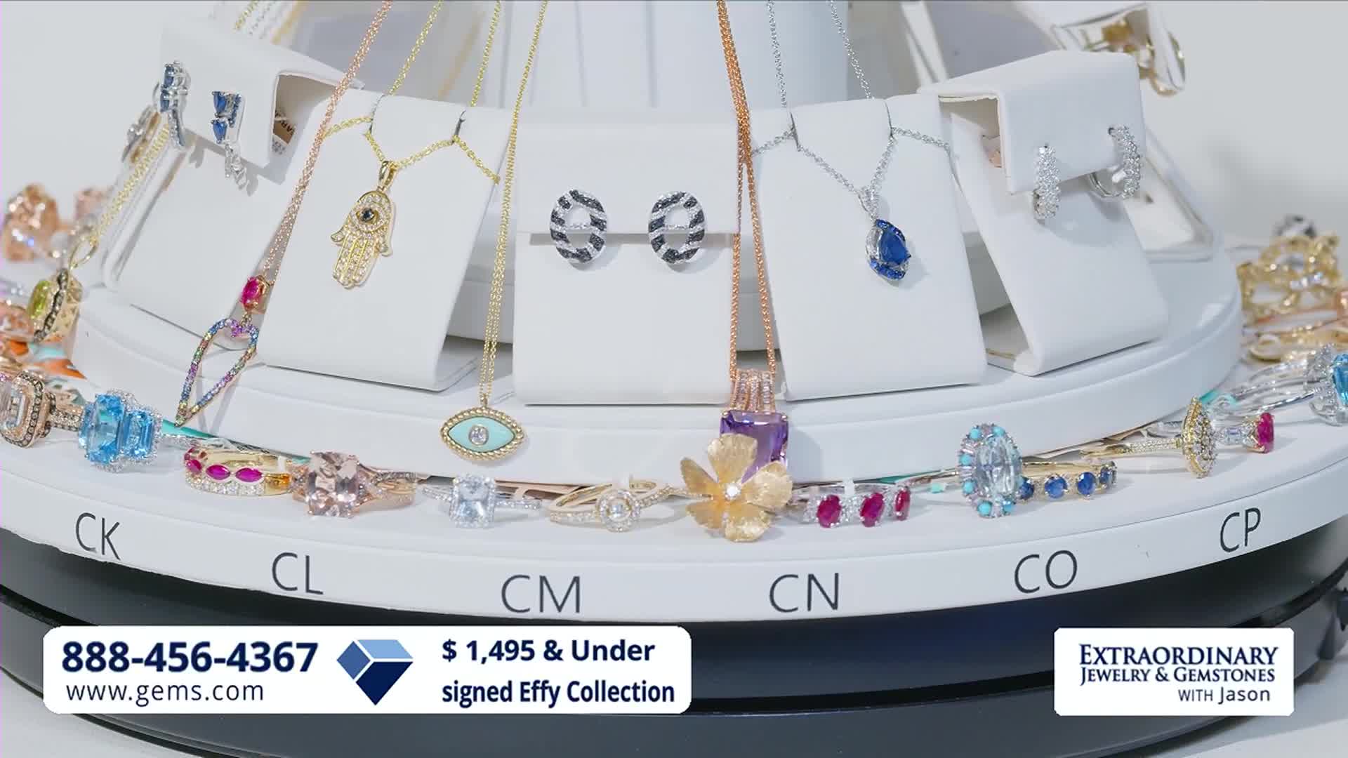 The jewelry display slowly rotates, showcasing necklaces and earrings on white stands. Rings with various gemstones are arranged on the white platform below.