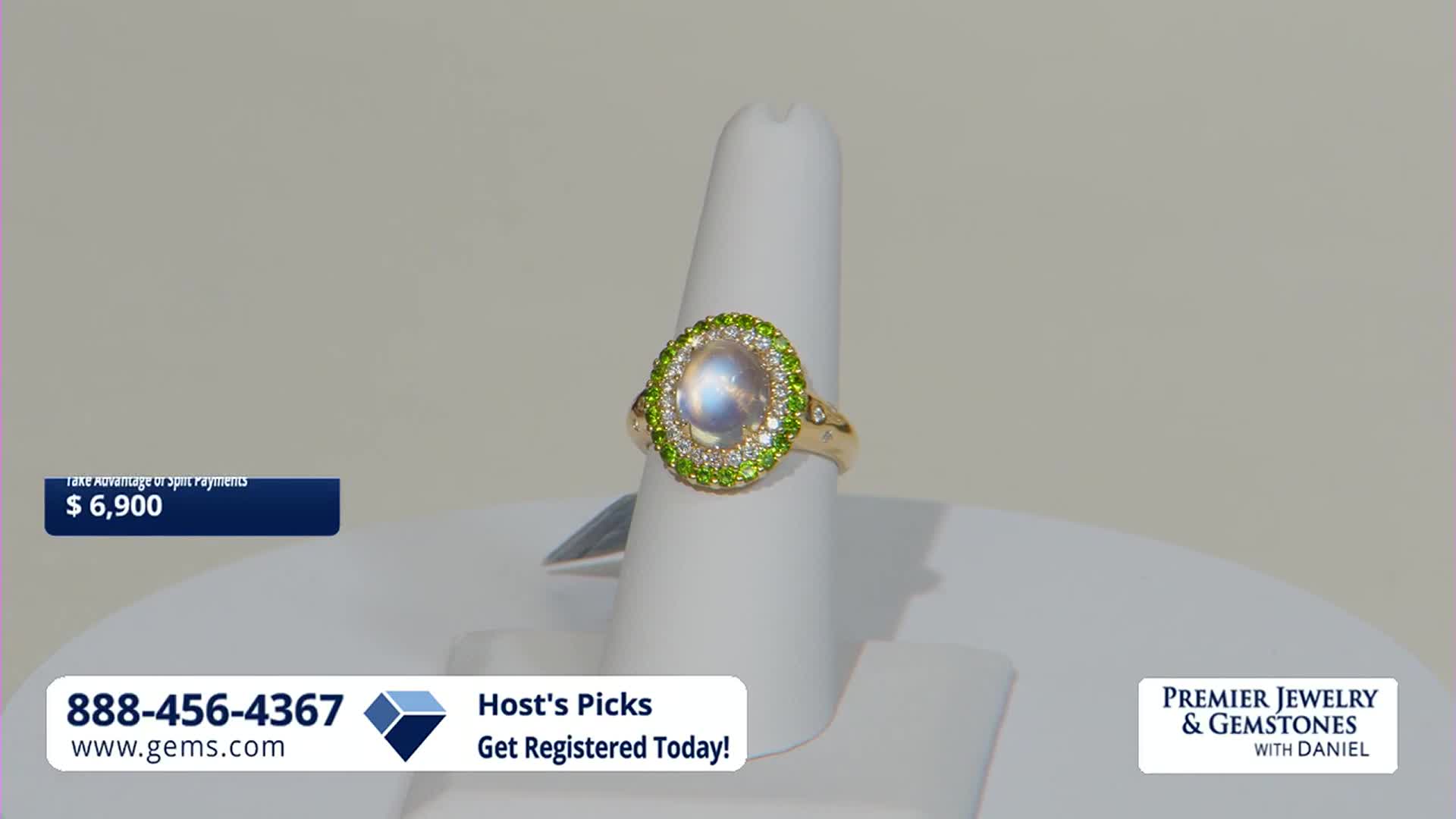 A gold ring with a large, milky stone and a halo of green gems sits on a white display. The price, $6,900, and a phone number for Gem Shopping Network are visible.