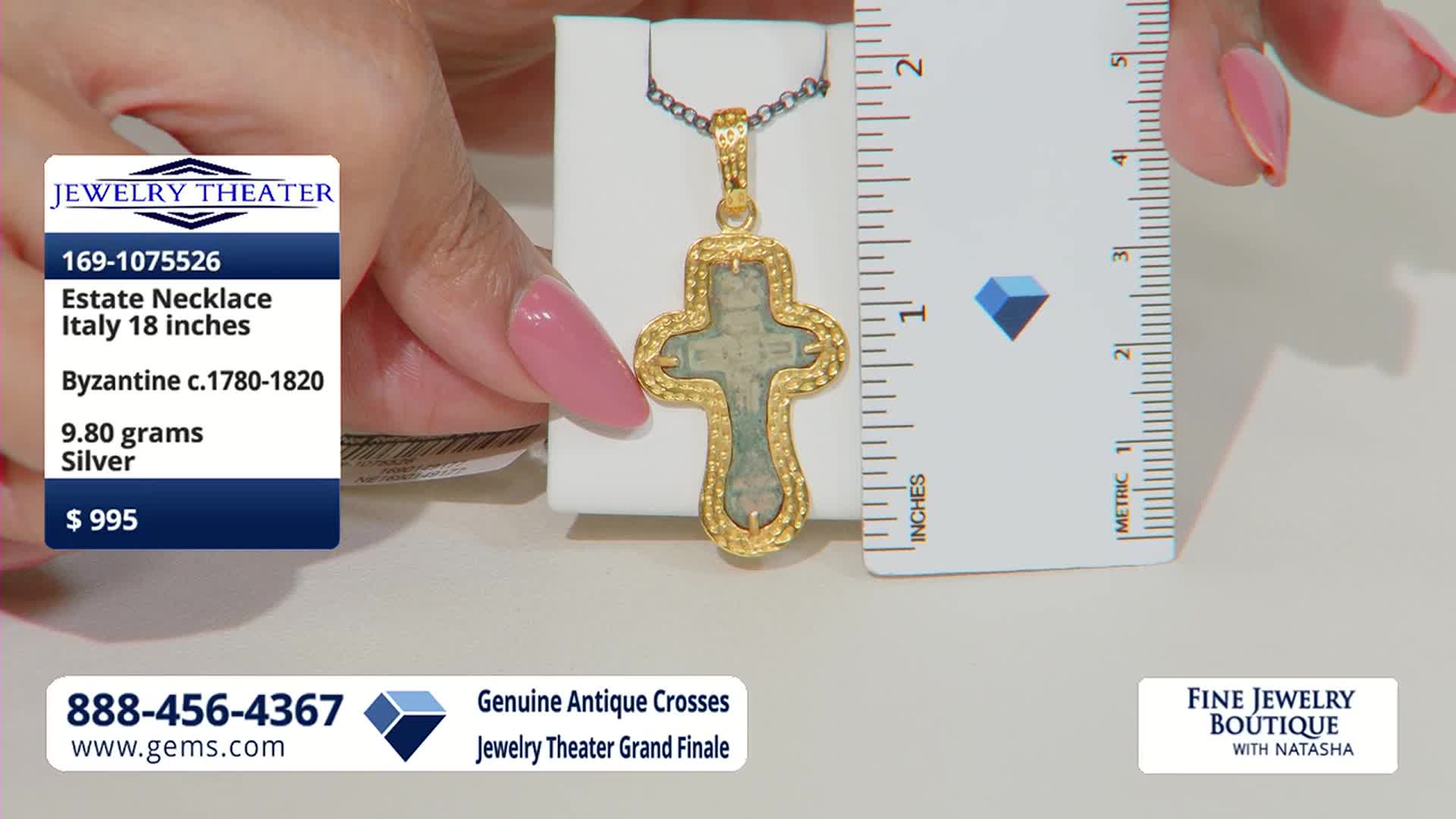 A hand with a pink-lacquered nail holds a ruler next to an ornate gold cross pendant. The pendant, featuring a pale green inset, rests on a white display stand.