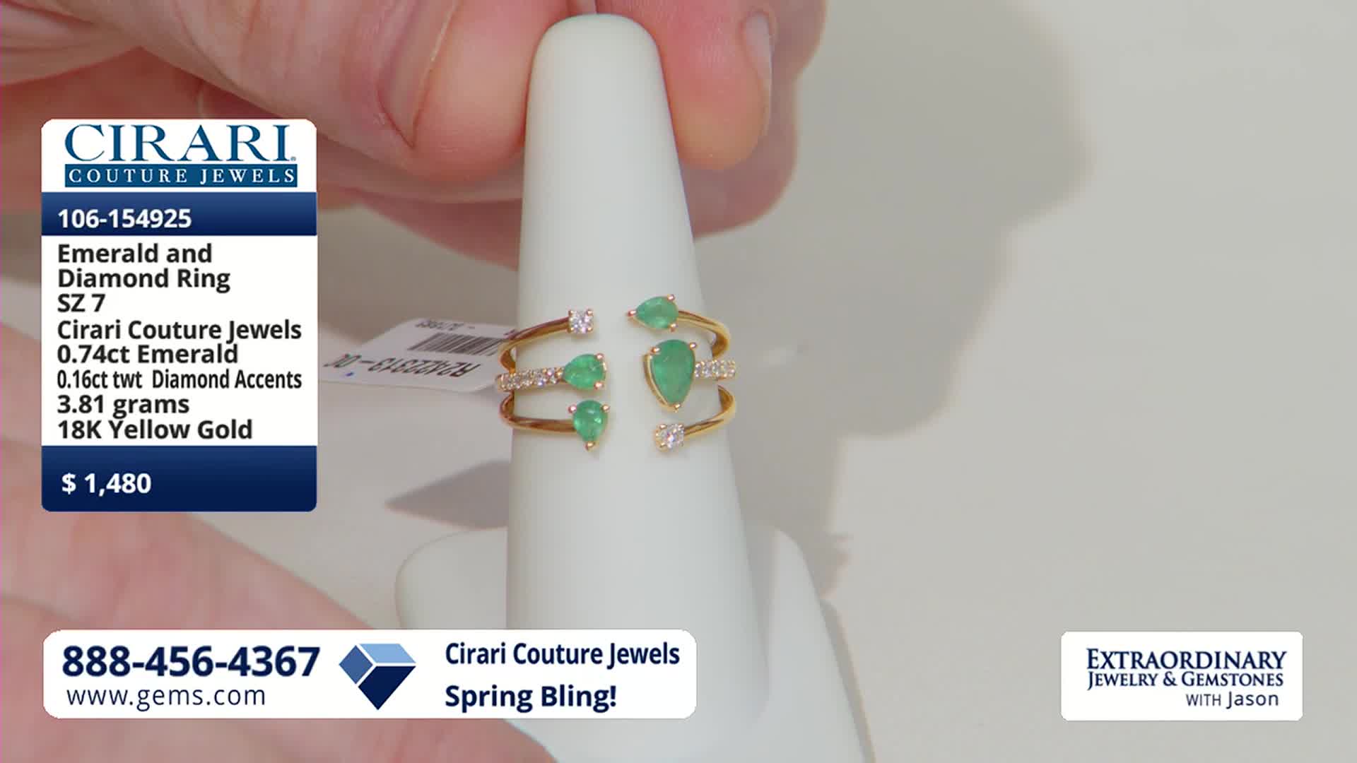 A hand is adjusting a gold ring on a white display. The ring features several pear-shaped emeralds and small diamonds.