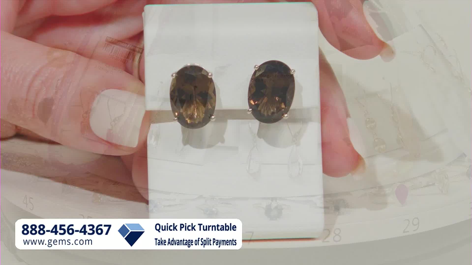 A hand gently adjusts a display stand holding a pair of oval, smoky quartz earrings. The Gem Shopping Network's logo and contact information appear at the bottom of the screen.