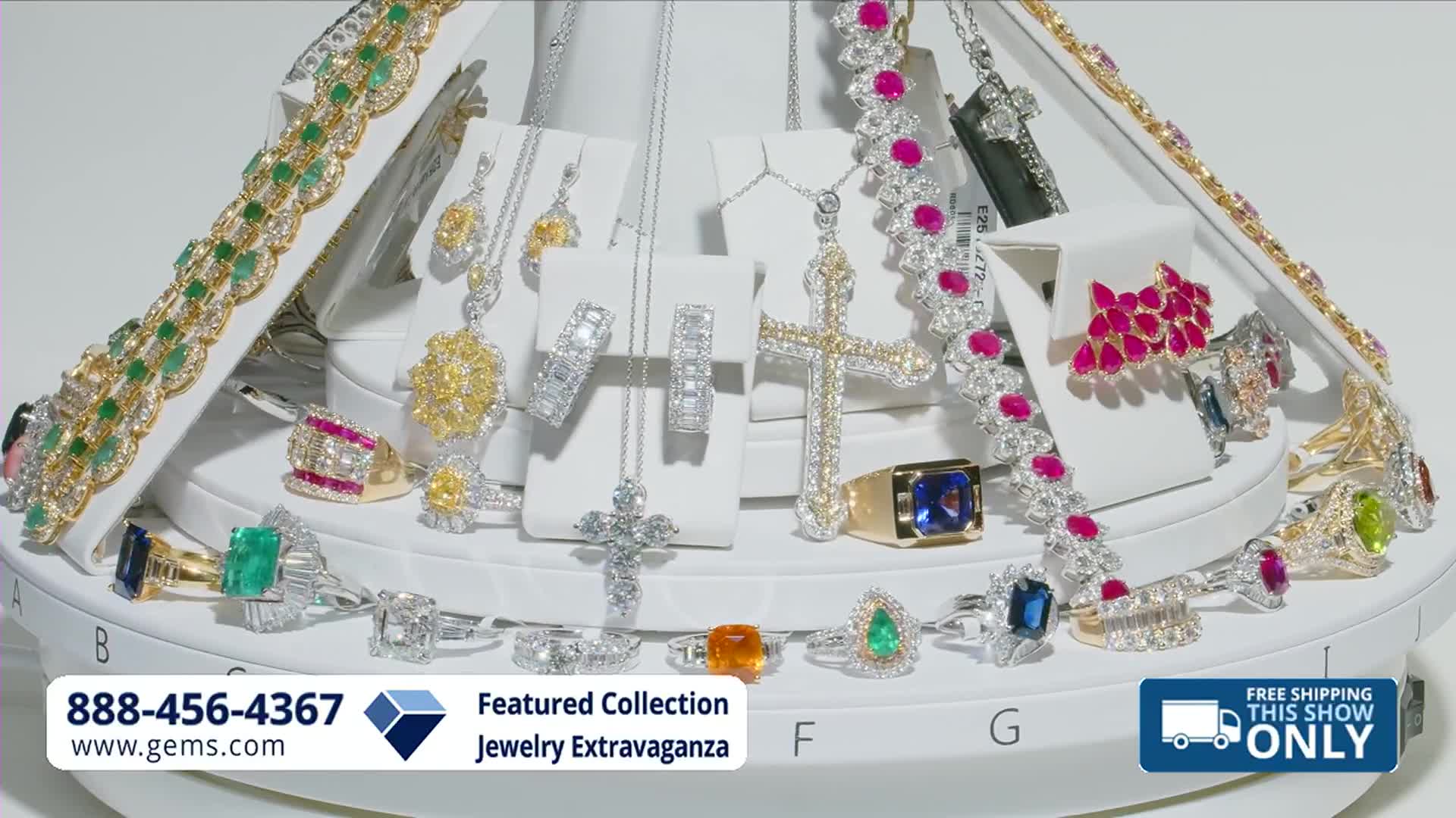 The display is slowly rotating, showcasing a dazzling array of diamond and gemstone jewelry. A gold ring with a large blue sapphire catches the light as it turns.