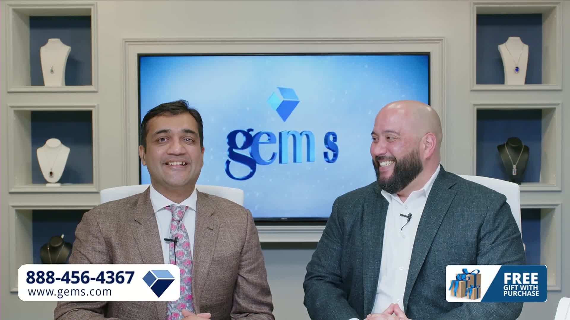 Two men smile as they present jewelry on a television screen. The Gem Shopping Network logo appears behind them, with a phone number and website at the bottom.