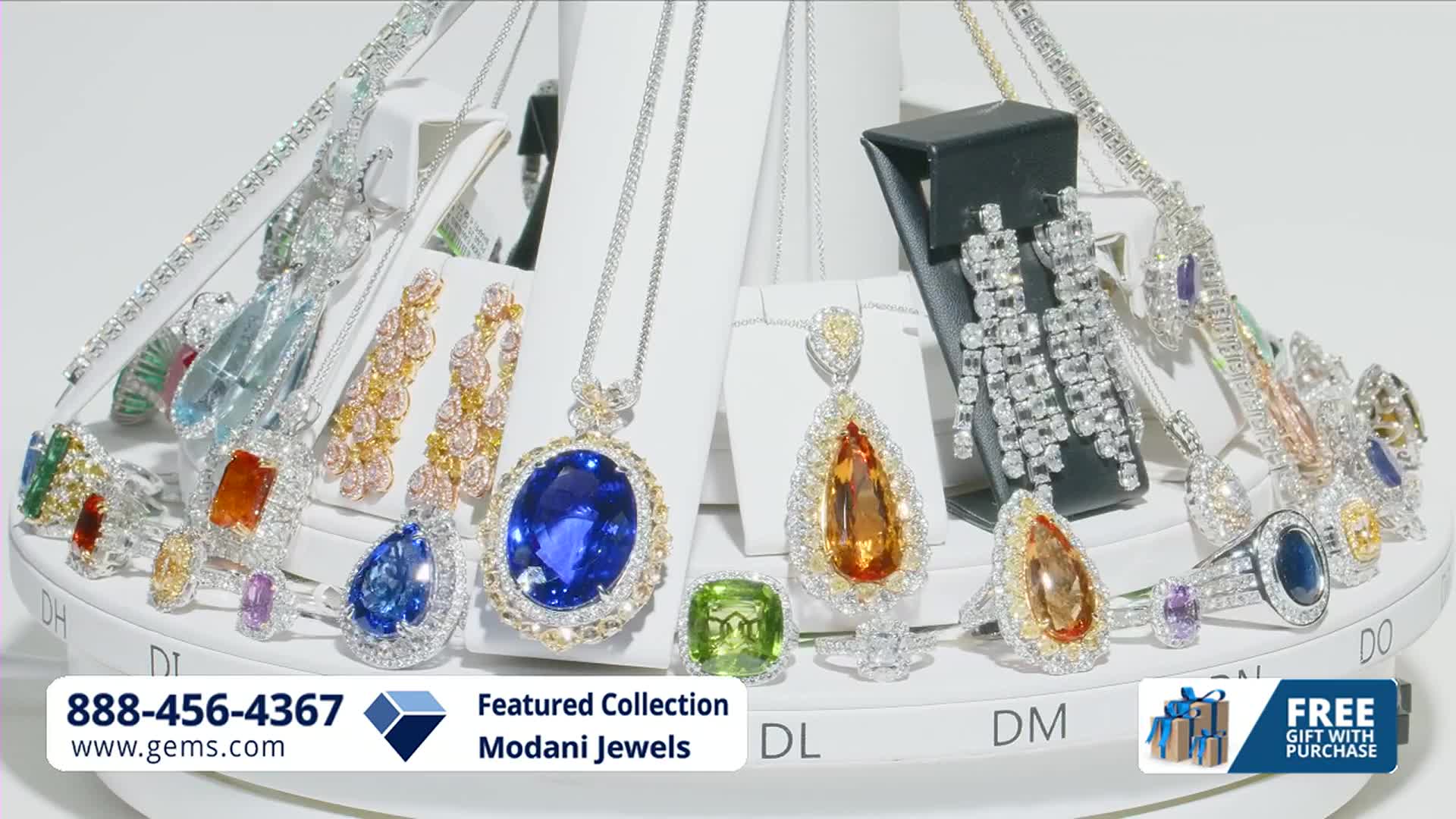 A display of sparkling necklaces, rings, and earrings, featuring large, colorful gemstones, rotates slowly. The Gem Shopping Network's featured collection, Modani Jewels, is presented with a phone number and website.