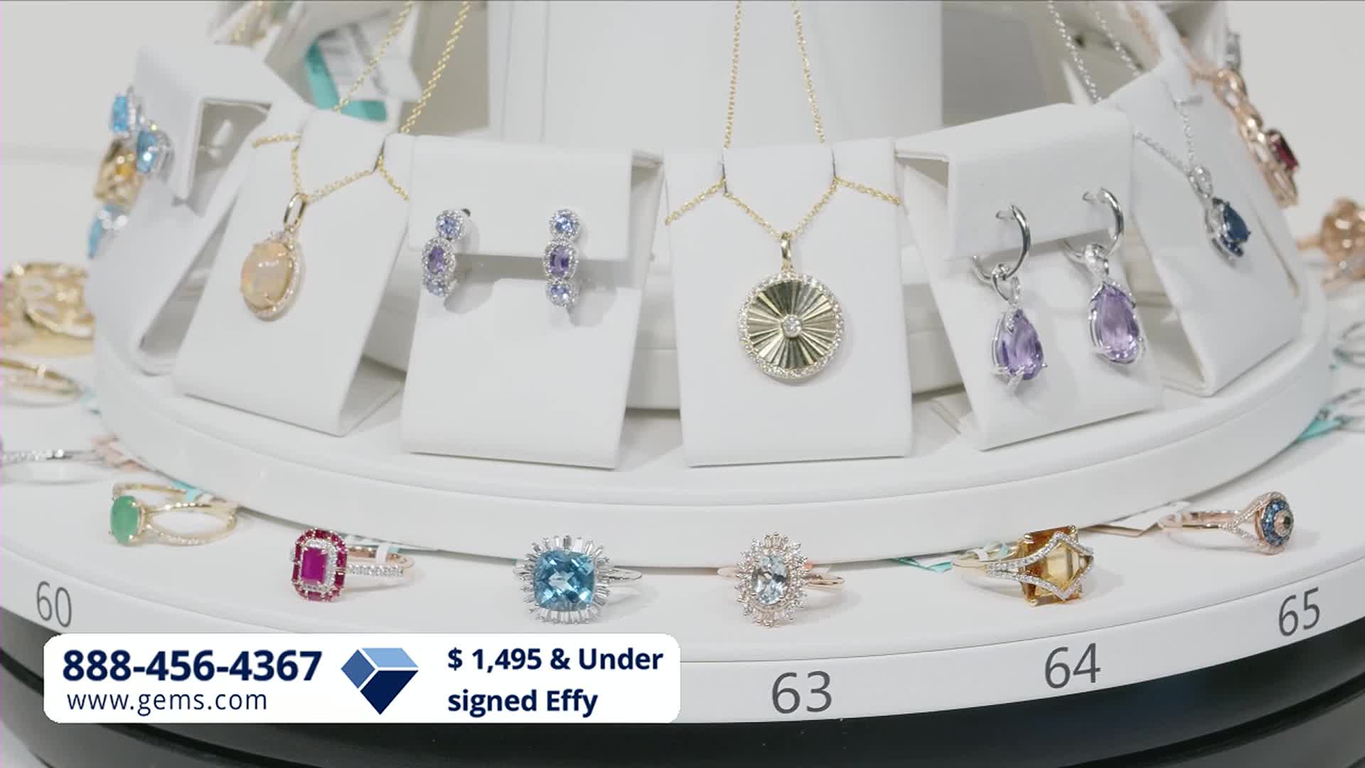The display is slowly turning, showcasing necklaces and earrings with colorful gemstones. Rings with large, faceted stones are arranged on the lower tier.