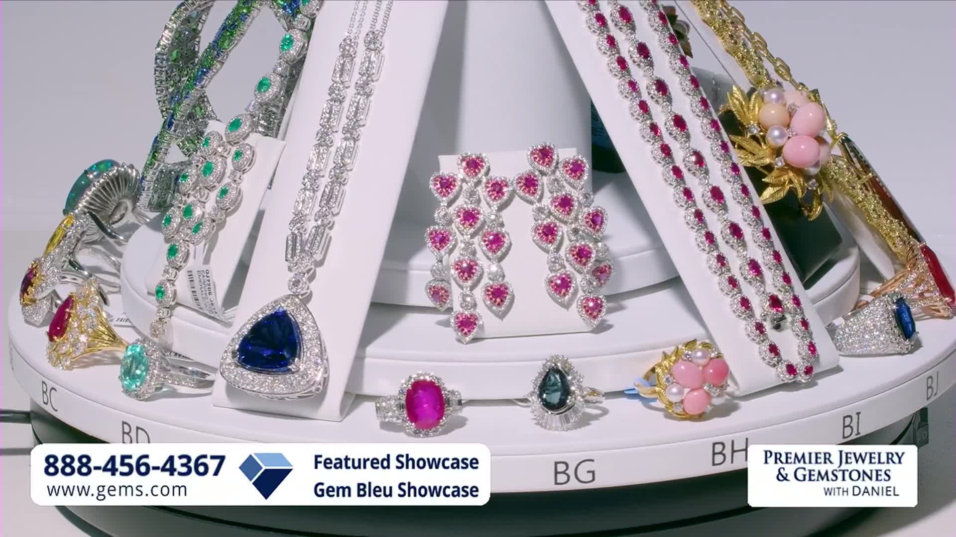 The jewelry display is slowly rotating, showcasing a variety of sparkling necklaces, rings, and earrings. A large blue sapphire pendant necklace catches the light as it turns.