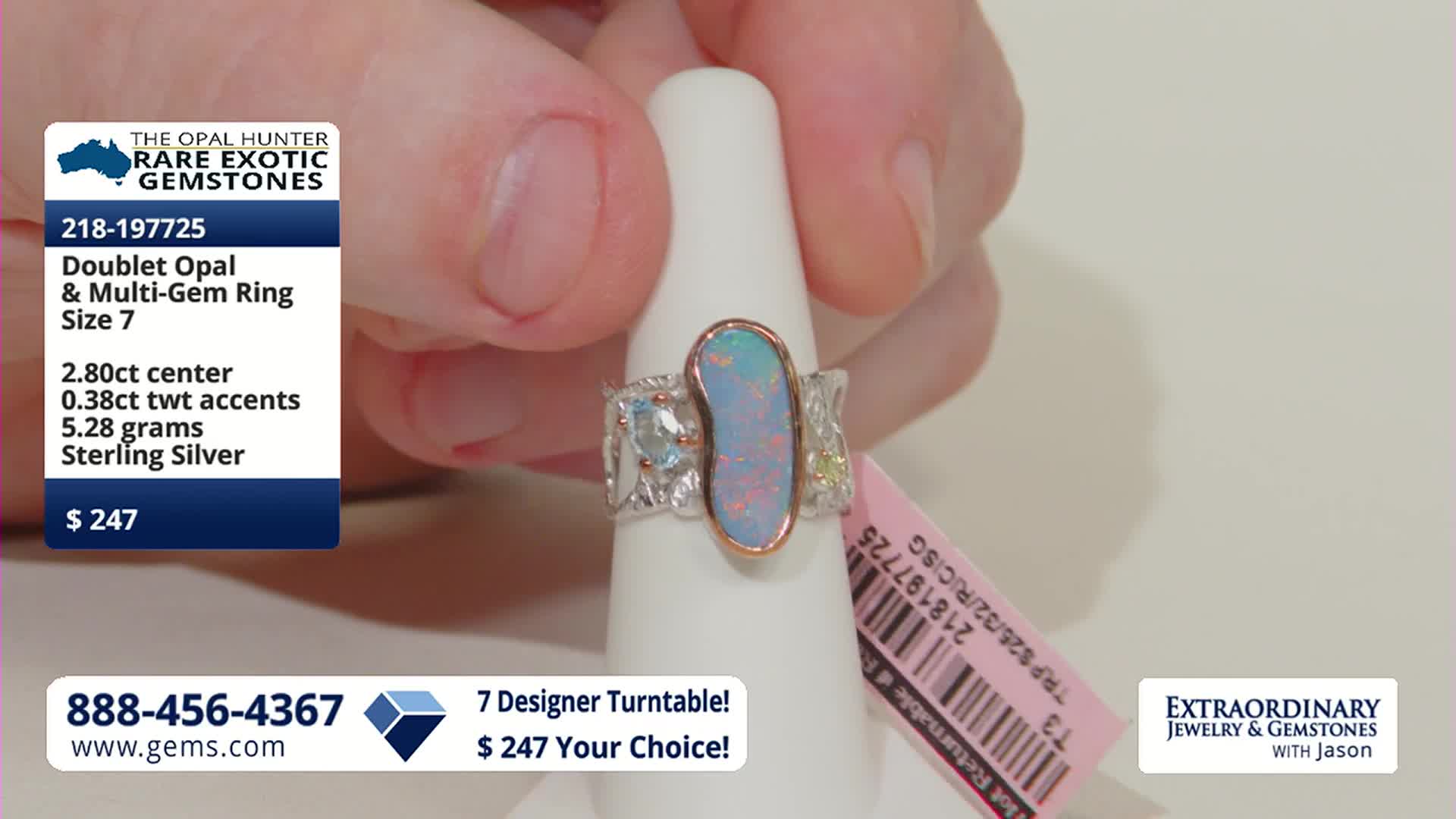 A hand holds a ring with a pale blue and pink opal center, flanked by smaller stones on a white display. The ring's band features a textured, silver finish.