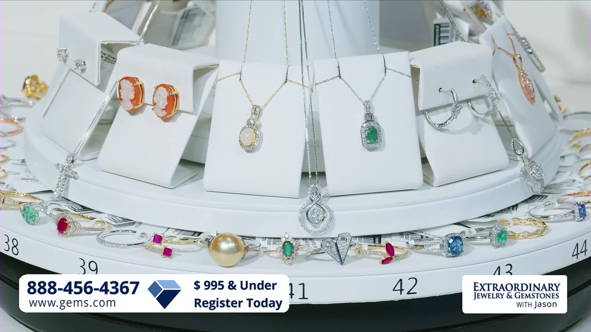 A display of necklaces and earrings rotates, showcasing precious stones. Rings with various gems line the base of the stand, each numbered for easy reference.