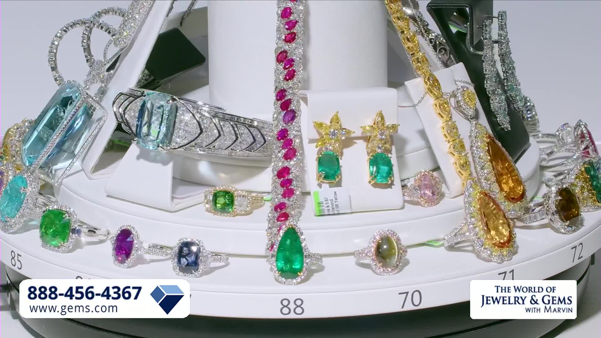 The display is slowly rotating, showcasing a collection of sparkling rings, bracelets, and earrings. A large emerald pendant hangs prominently from a ruby-studded bracelet.