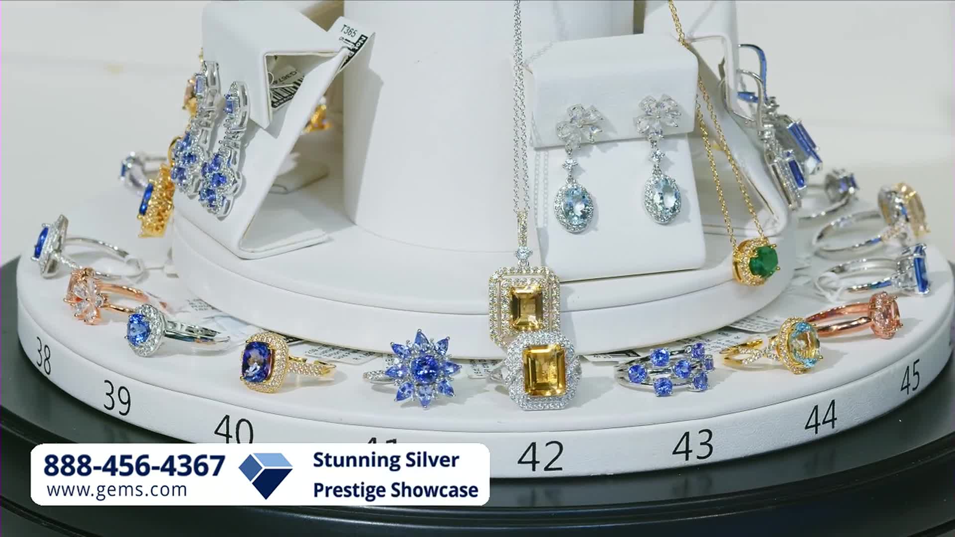 A display of rings and necklaces with colorful gemstones sits on a tiered stand. The Gem Shopping Network logo and a phone number are visible at the bottom.
