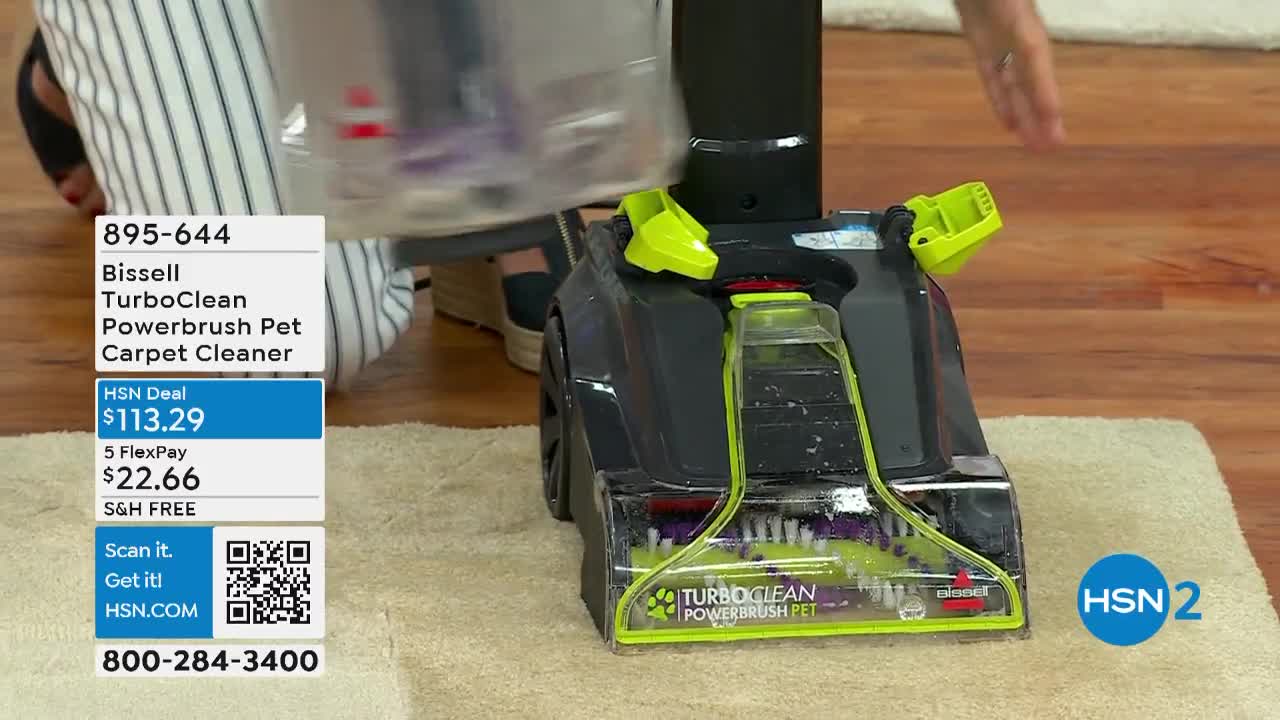 A carpet cleaner, labeled "TurboClean Powerbrush Pet," sits on a light-colored rug. The presenter holds the clear water tank above the machine on the set of HSN2.
