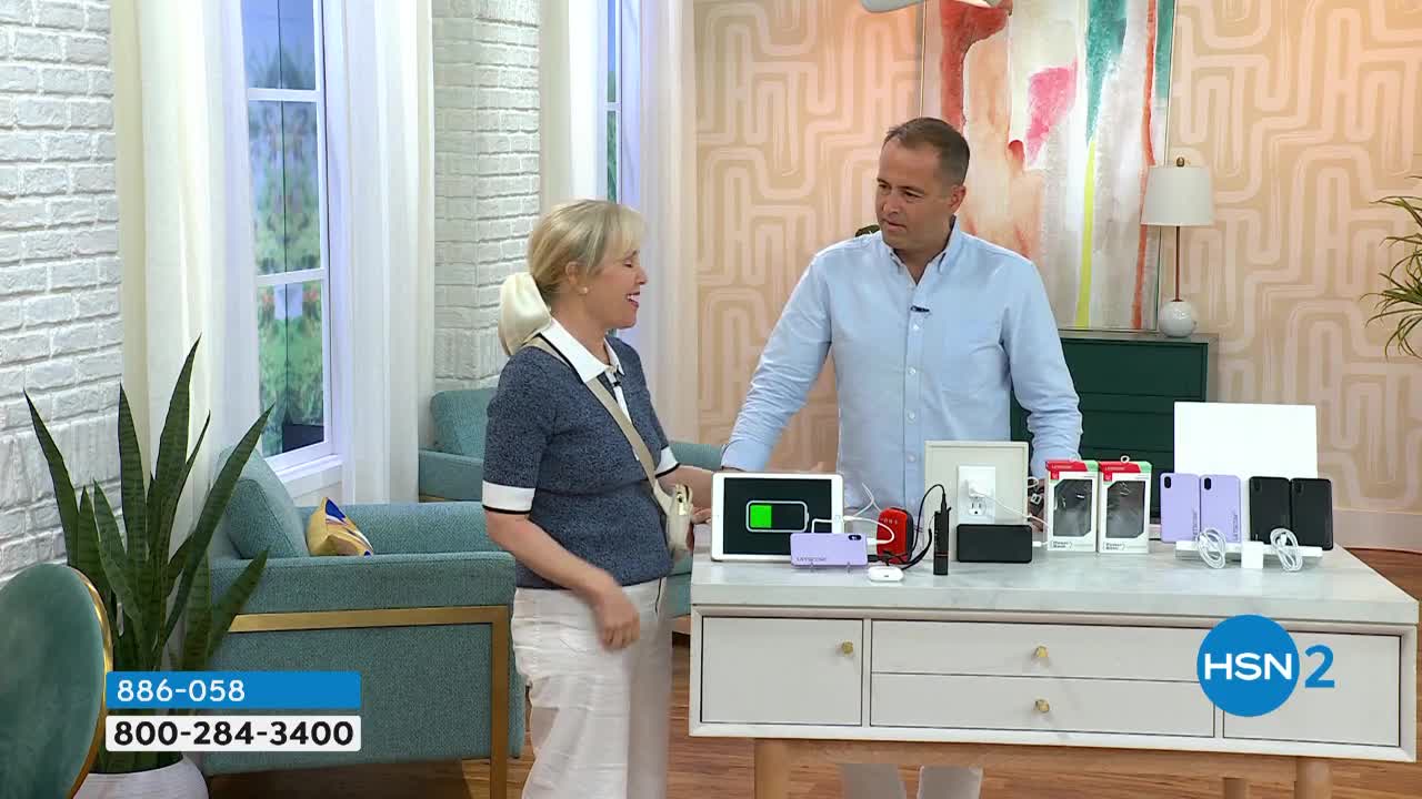 A woman in a blue sweater and white pants gestures towards a table displaying various tech accessories. The man beside her, in a light blue shirt, looks on as they present the products on the HSN2 set.
