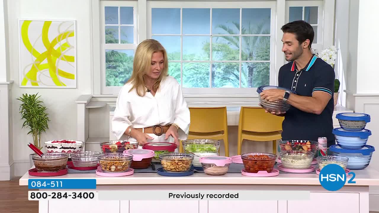 The man holds up a clear bowl filled with food, gesturing toward the woman beside him. On the table, various glass containers with colorful lids and contents are arranged for the HSN2 presentation.
