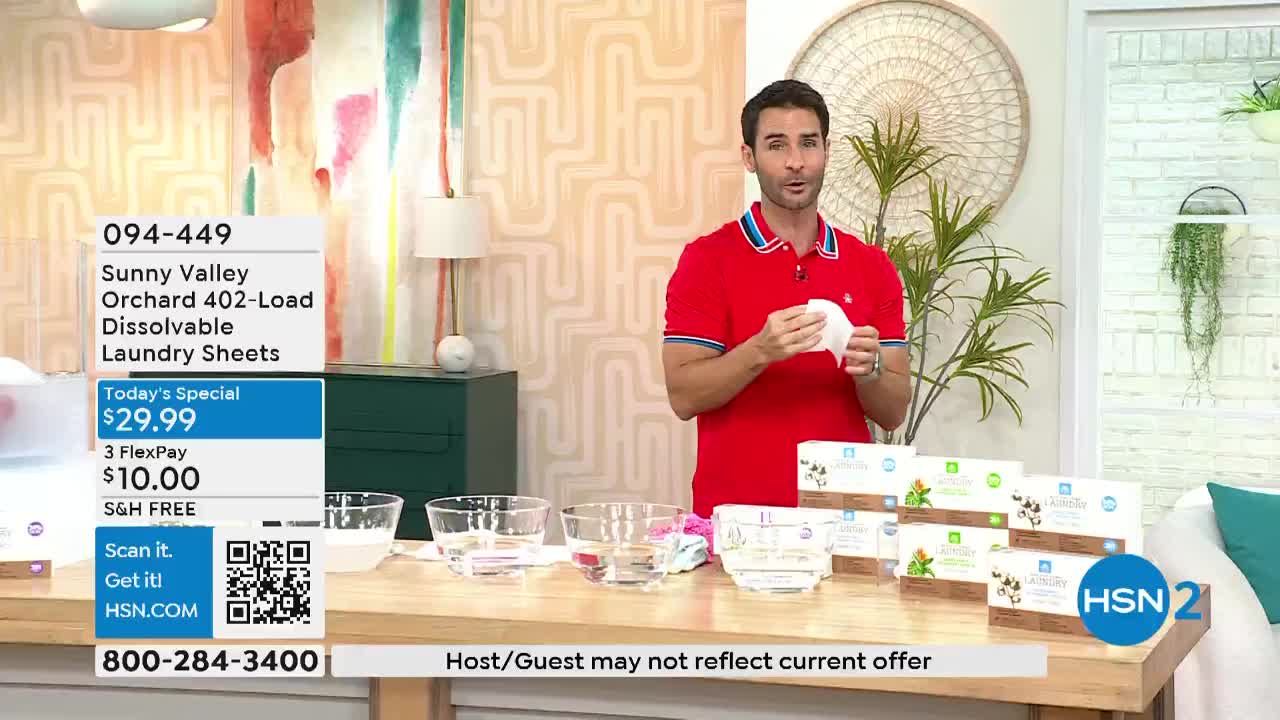 A man in a red polo shirt holds up a white laundry sheet, demonstrating its use. Several clear bowls of water sit on a table in front of him, along with boxes of the product.
