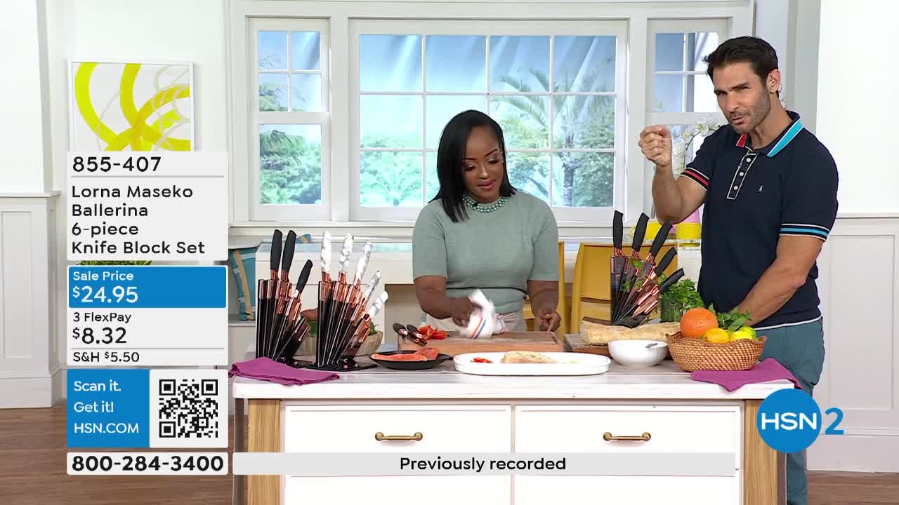 A woman wipes a cutting board while the man to her right gestures with his finger. Behind them, various knives are displayed on the counter, alongside a basket of fruit.
