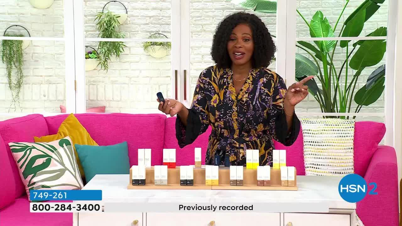 A woman in a patterned robe gestures towards several small, white cosmetic product boxes on a wooden display. She holds a small, dark object in her other hand as she speaks, seemingly presenting the items on HSN2.

