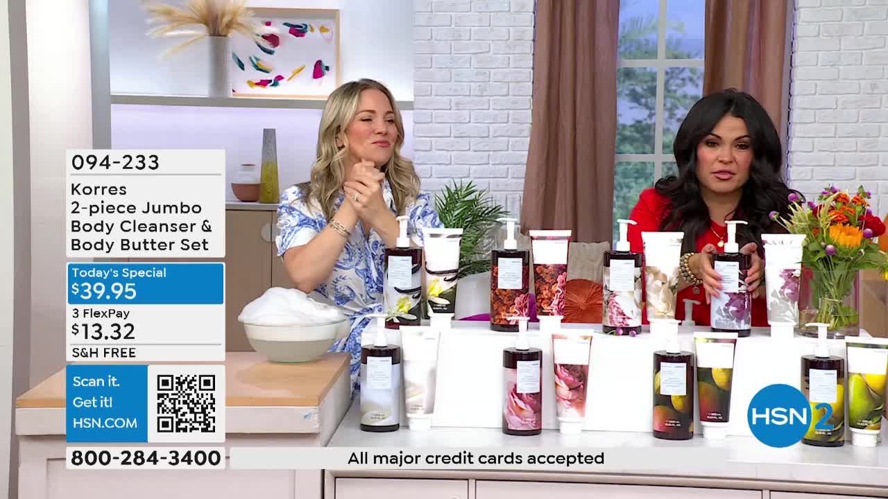 Two women on HSN2 stand behind a table displaying several bottles of body products. One woman gestures with her hands, while the other holds up a bottle, likely describing its features.
